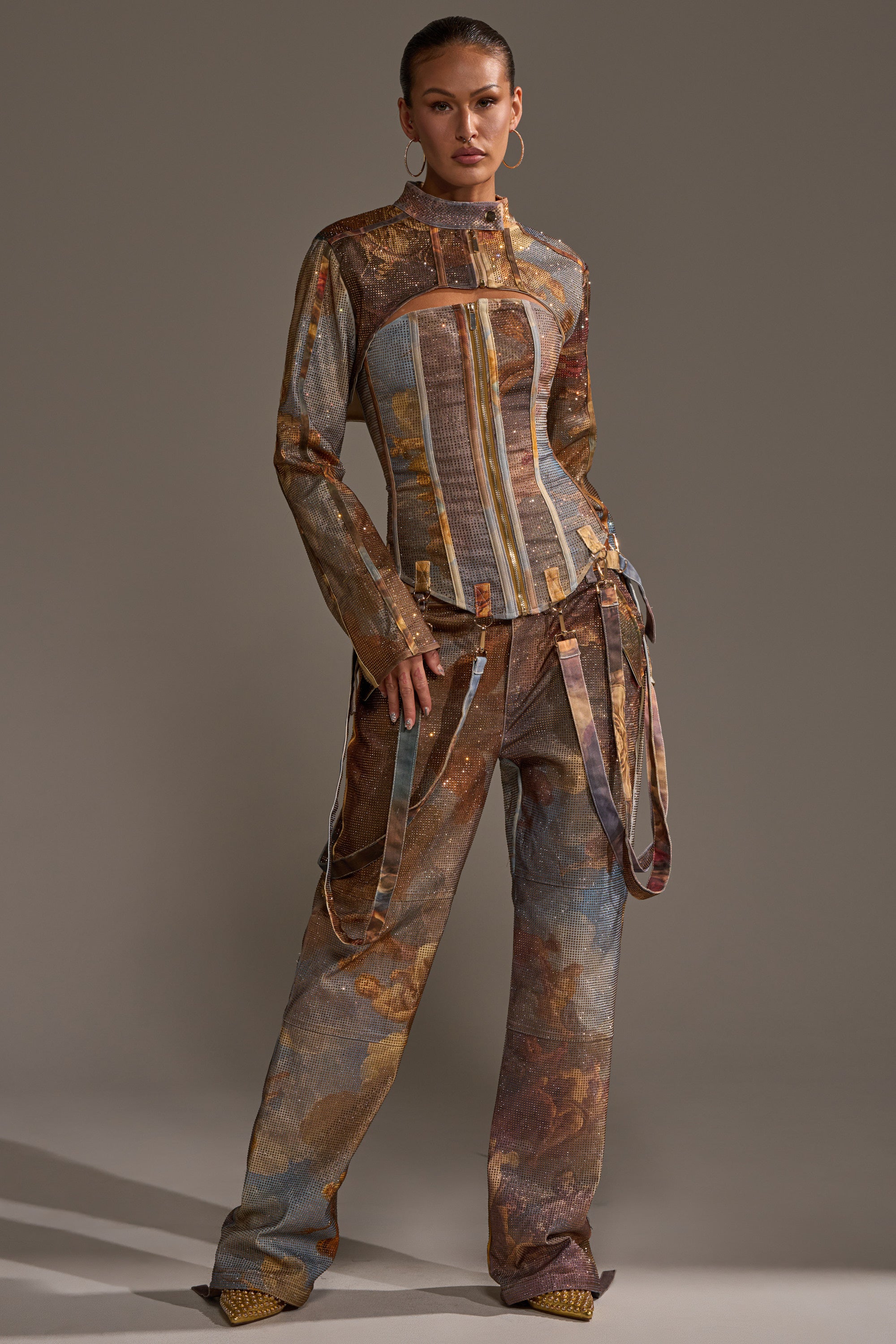 A woman exudes confidence in the IN THE FUTURE RHINESTONE BOLERO, styled with matching wide-leg pants featuring strap details. Her hair is pulled back, complemented by large hoop earrings and textured pointed shoes.