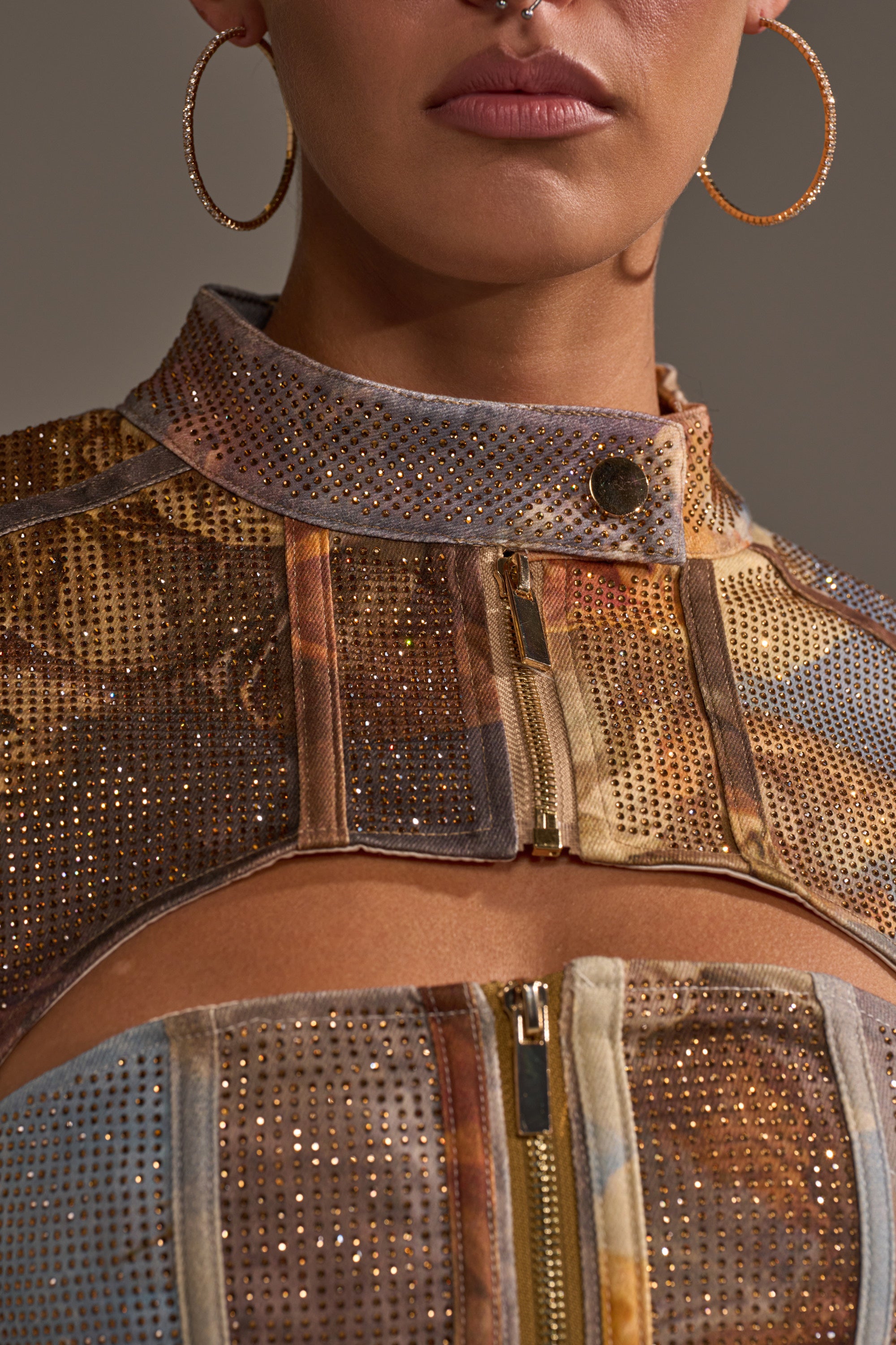 Close-up of a person wearing the IN THE FUTURE RHINESTONE BOLERO, featuring a multicolored graphic print with gold studs and zippers. Large hoop earrings, a septum piercing, and glossy lips complete the look.