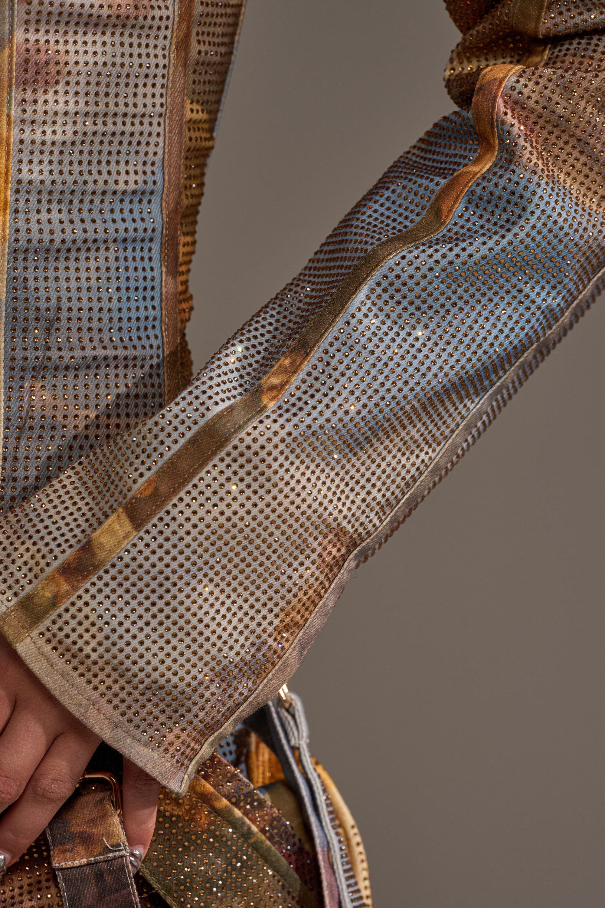 Close-up of a person wearing the IN THE FUTURE RHINESTONE BOLERO—a long-sleeved, metallic mesh-textured cropped jacket featuring a multicolored patterned design with small reflective dots and blended neutral tones.