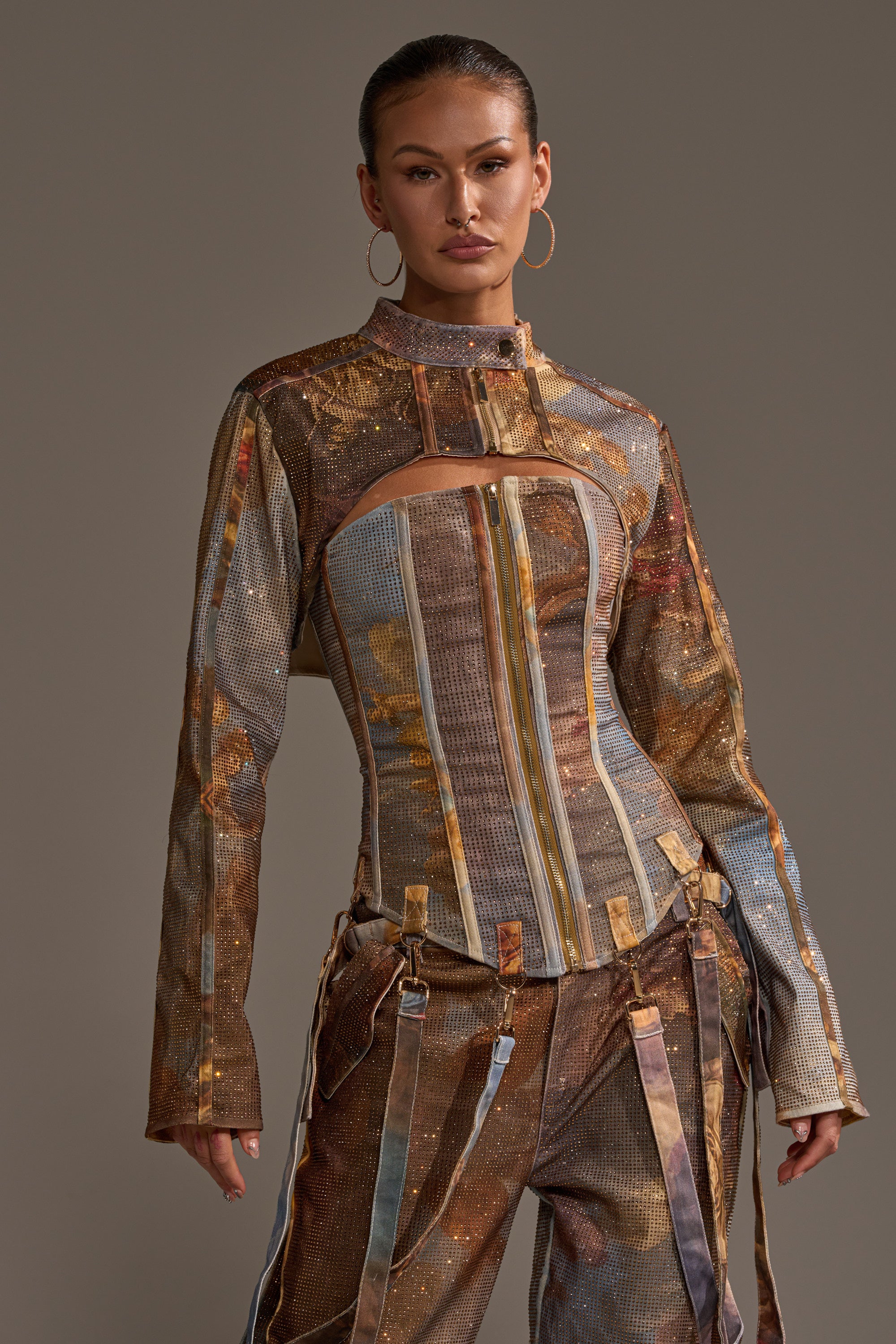 A woman wears the IN THE FUTURE RHINESTONE BOLERO over a structured, multicolored metallic outfit with a high collar, chest cutout, vertical stripes, and suspenders, paired with matching pants, hoop earrings, and her hair pulled back.