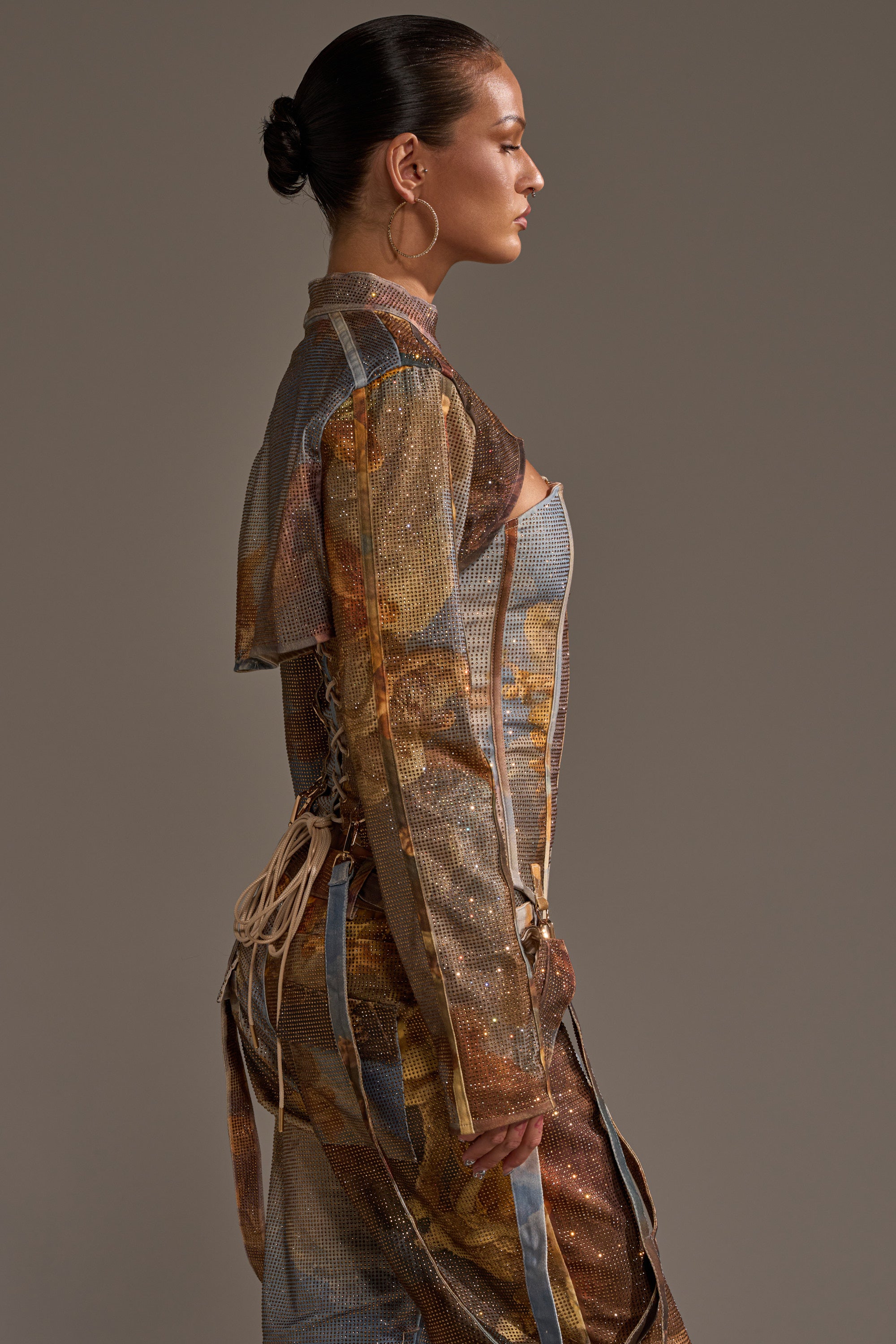A woman with slicked-back hair in a bun models the IN THE FUTURE RHINESTONE BOLERO, an earth-toned piece with metallic details and bold graphic prints, posing in profile against a plain background.