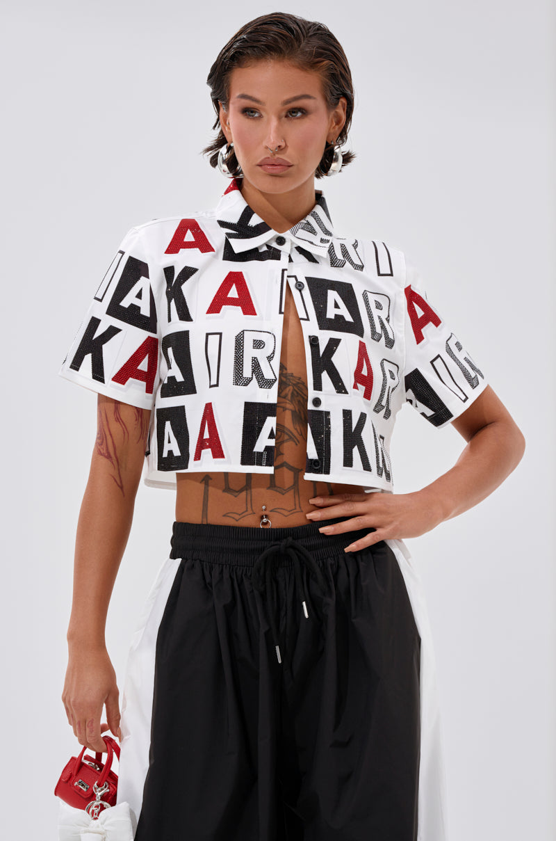 Someone with short dark hair wears the READ IT BABY BUTTON DOWN by AKIRA Label, a white cropped shirt with bold black and red rhinestone lettering spelling “AIR” and “A.” They pair it with black pants and a small red accessory on a light background. 0