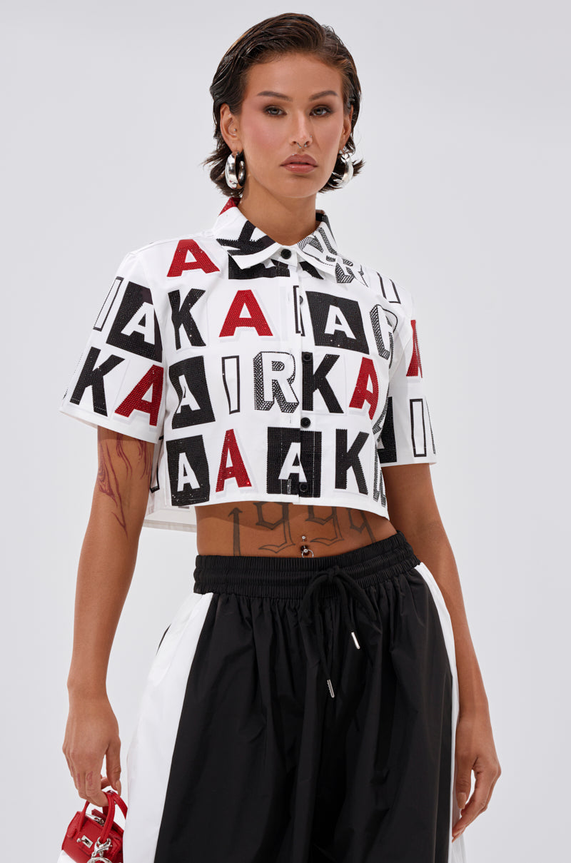 A person with short, slicked-back hair wears the READ IT BABY BUTTON DOWN featuring bold black and red letter patterns, stylish black and white wide-leg pants, statement earrings, and carries a small red handbag. 1