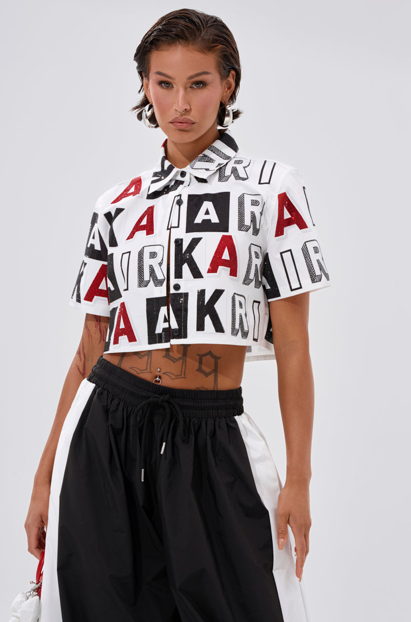 A woman stands confidently against a neutral background, wearing the READ IT BABY BUTTON DOWN with bold black and red "AKIR" lettering, paired with loose black and white pants. 2