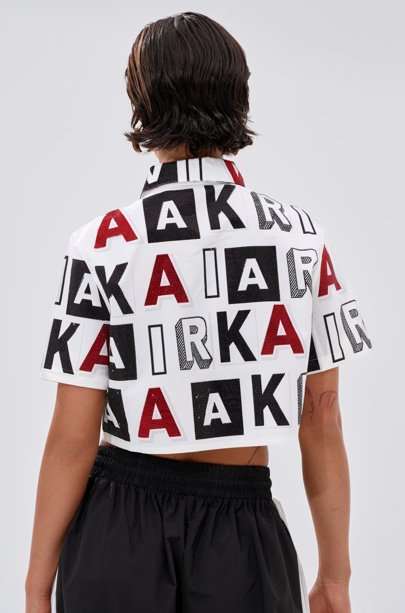 A person with short, wet-look hair wears the white READ IT BABY BUTTON DOWN featuring bold black and red “A” and “K” square patterns, paired with black pants. The outfit is shown from the back. 4