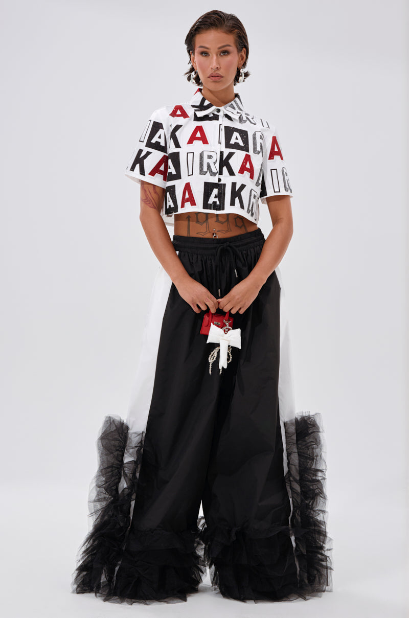 A woman stands against a plain background wearing the READ IT BABY BUTTON DOWN with rhinestone lettering, paired with wide black pants featuring tulle ruffles at the hem and a decorative waist tie. 5