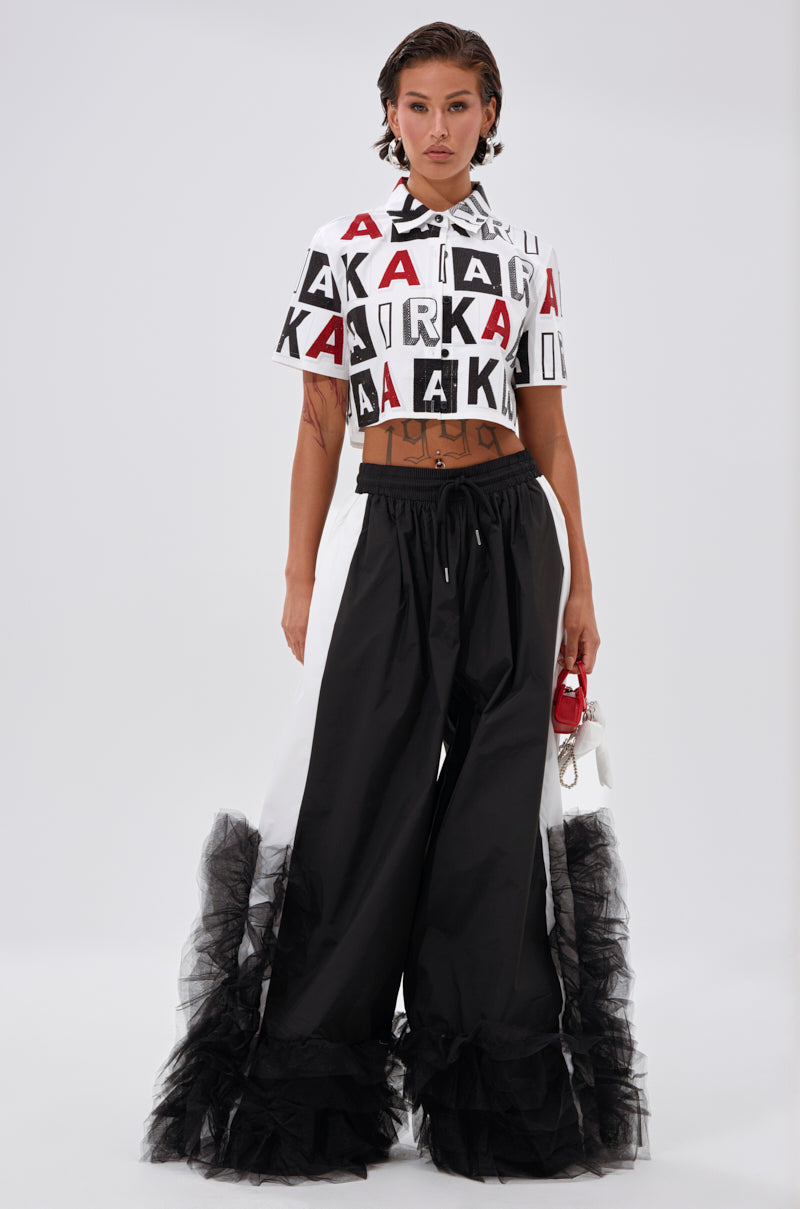 A person stands against a plain background wearing the AKIRA Label READ IT BABY BUTTON DOWN, a cropped white top with rhinestone block lettering, paired with wide black pants featuring sheer ruffles. They hold a small red and white accessory. 6