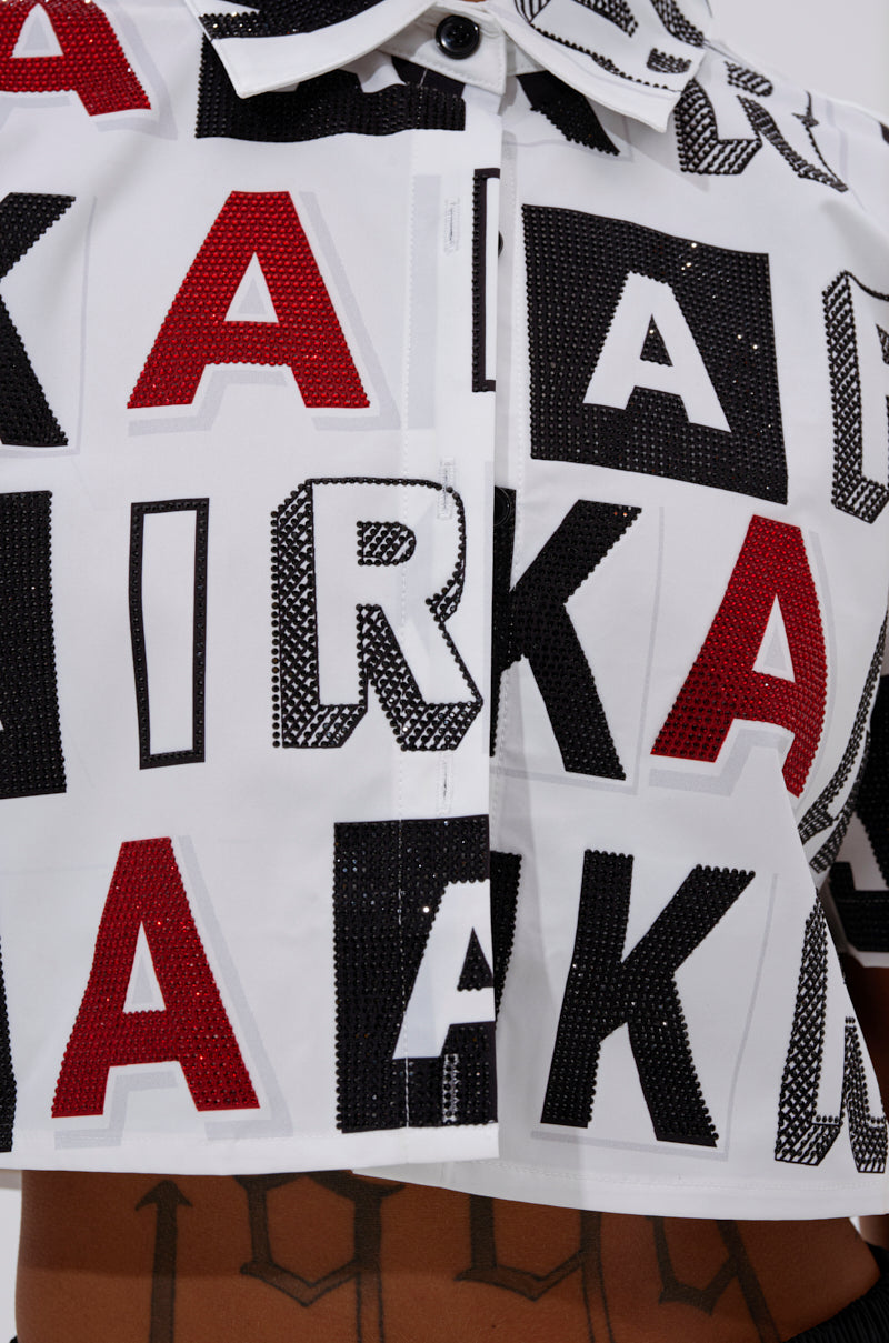 Close-up of the READ IT BABY BUTTON DOWN by AKIRA Label, a cropped white shirt with bold rhinestone "K" and "A" lettering, revealing part of a tattooed torso. 7