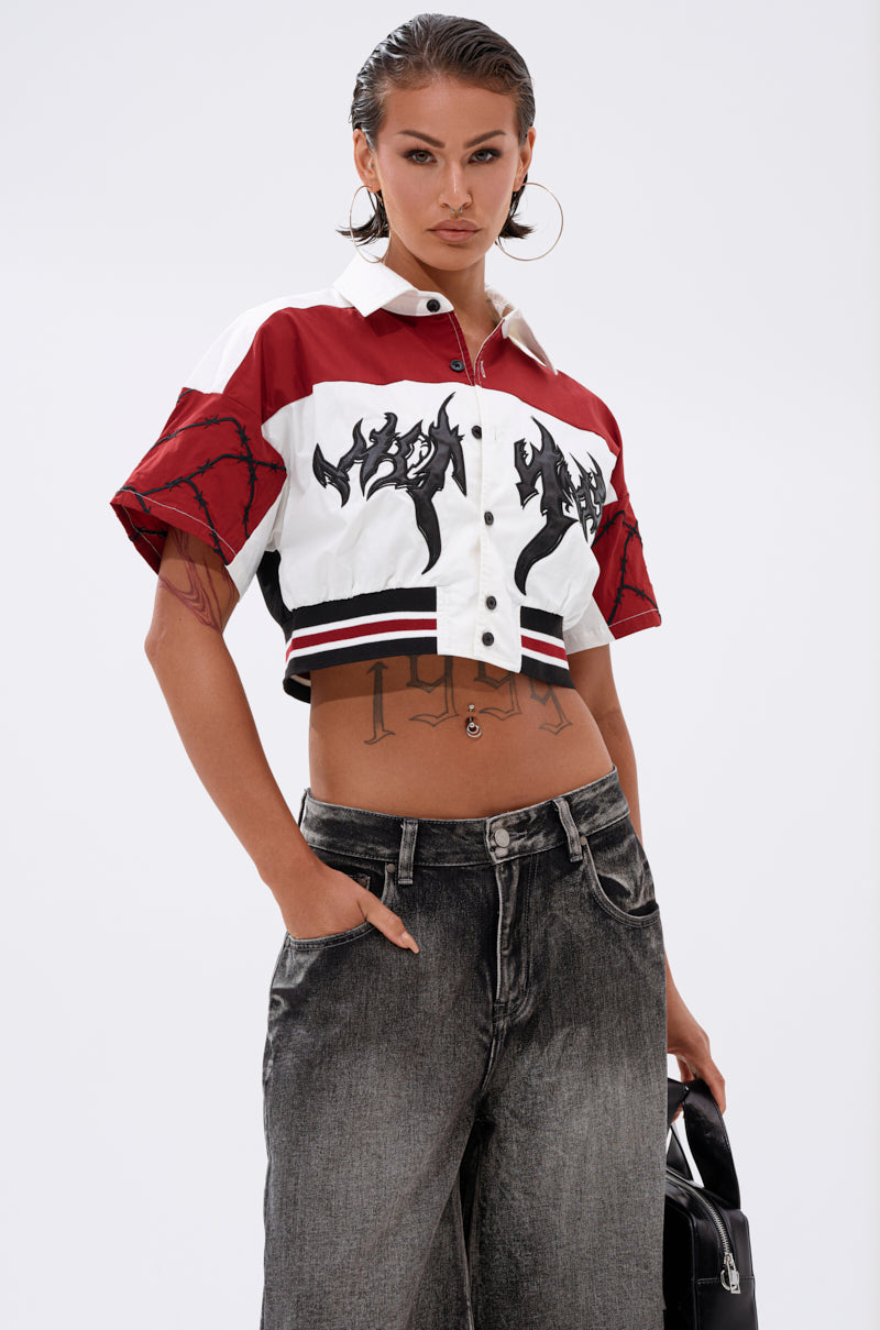 A person with slicked-back hair confidently poses with one hand in their pocket, wearing the WINNING TEAM BUTTON DOWN featuring bold graphics, loose dark jeans, large hoop earrings, and holding a black bag. 0