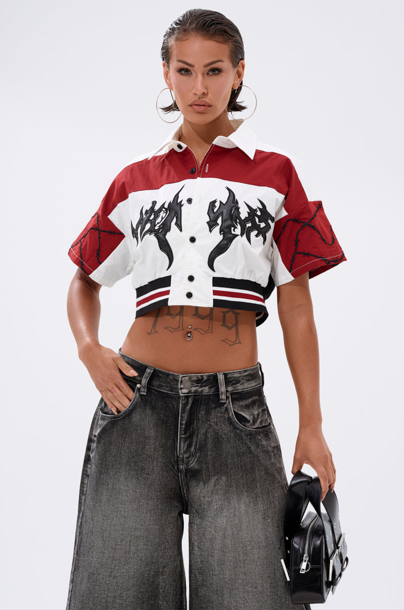 A woman styles the WINNING TEAM BUTTON DOWN, a red and white cotton boxy cropped shirt, with wide-leg faded jeans, large hoop earrings, slicked-back hair, visible torso tattoos, and a small black bag. 1