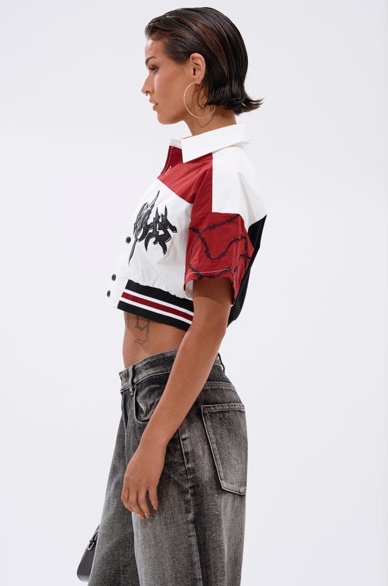 A person with short, slicked-back hair wears the WINNING TEAM BUTTON DOWN—a boxy cropped jacket with a red and white color block design, black graphic—paired with high-waisted gray jeans, shown in profile against a plain background. 2