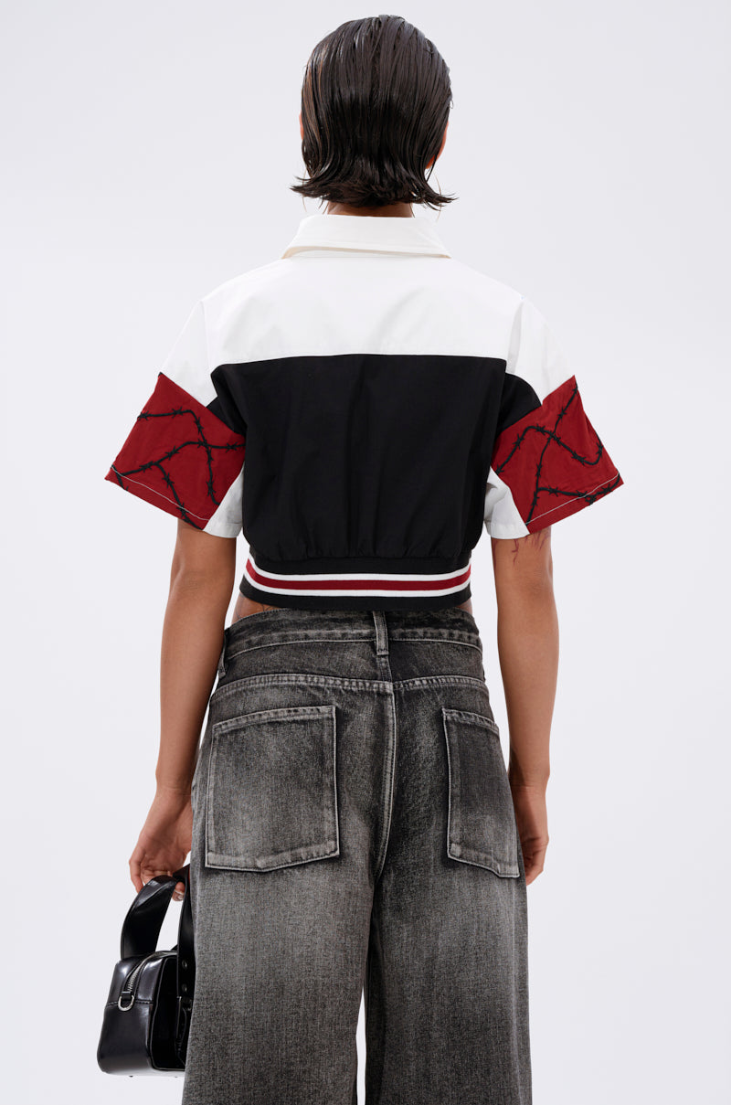 A person with wet, slicked-back hair is seen from the back, wearing the WINNING TEAM BUTTON DOWN in black, white, and red color blocks with wide-leg faded black jeans and holding a small black handbag. 3