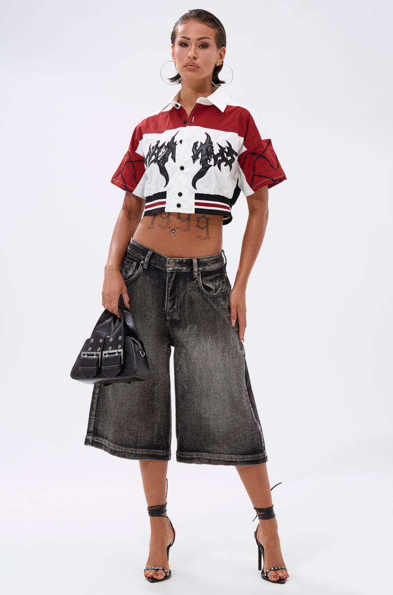 A person stands confidently in the WINNING TEAM BUTTON DOWN, loose dark denim knee-length shorts, black high heels, large hoop earrings, and holding a small black handbag. 5