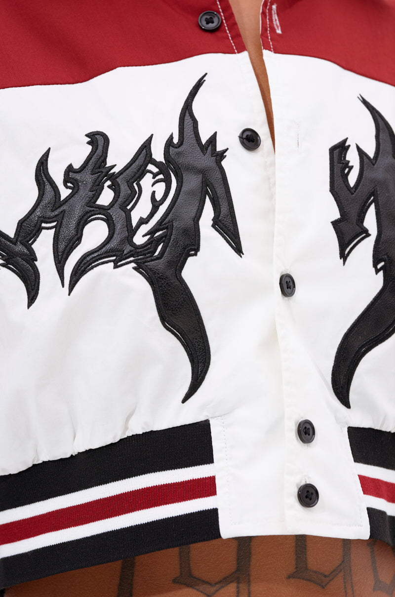 Close-up of the WINNING TEAM BUTTON DOWN—a boxy cropped jacket featuring black abstract designs on a red and white background with striped hems. A tattoo is partially visible on the wearer’s skin. 6