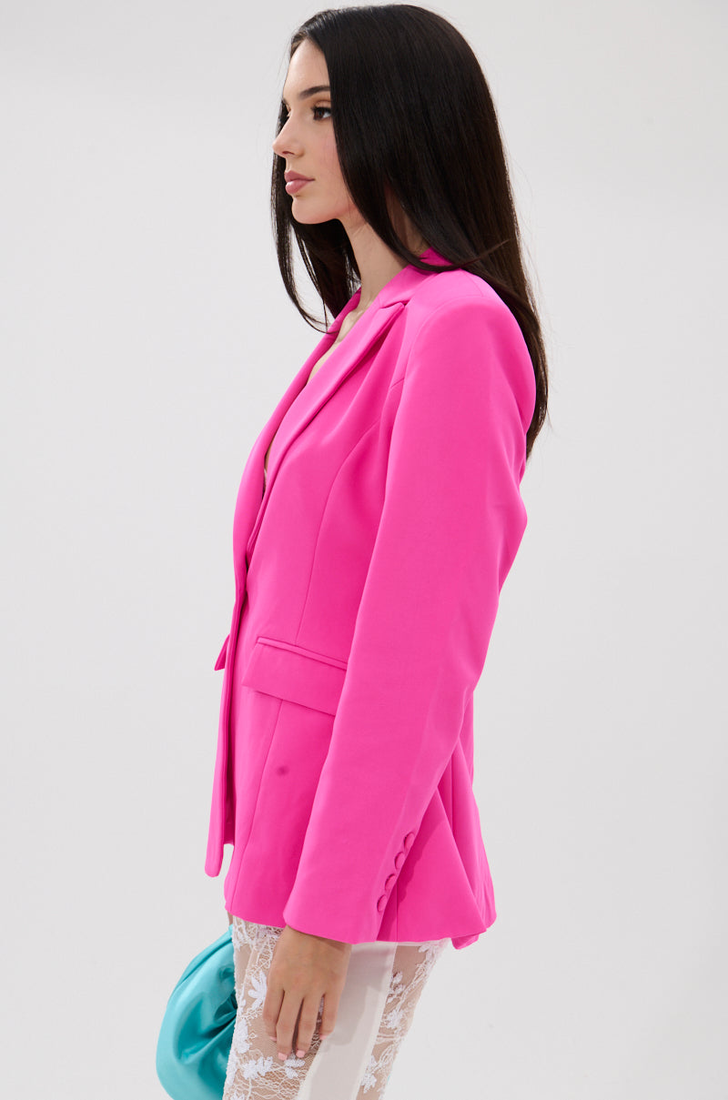 Woman with long dark hair wears the PRETTY IN PINK FITTED BLAZER and white lace pants, holding a turquoise clutch, posing against a plain white background. 3