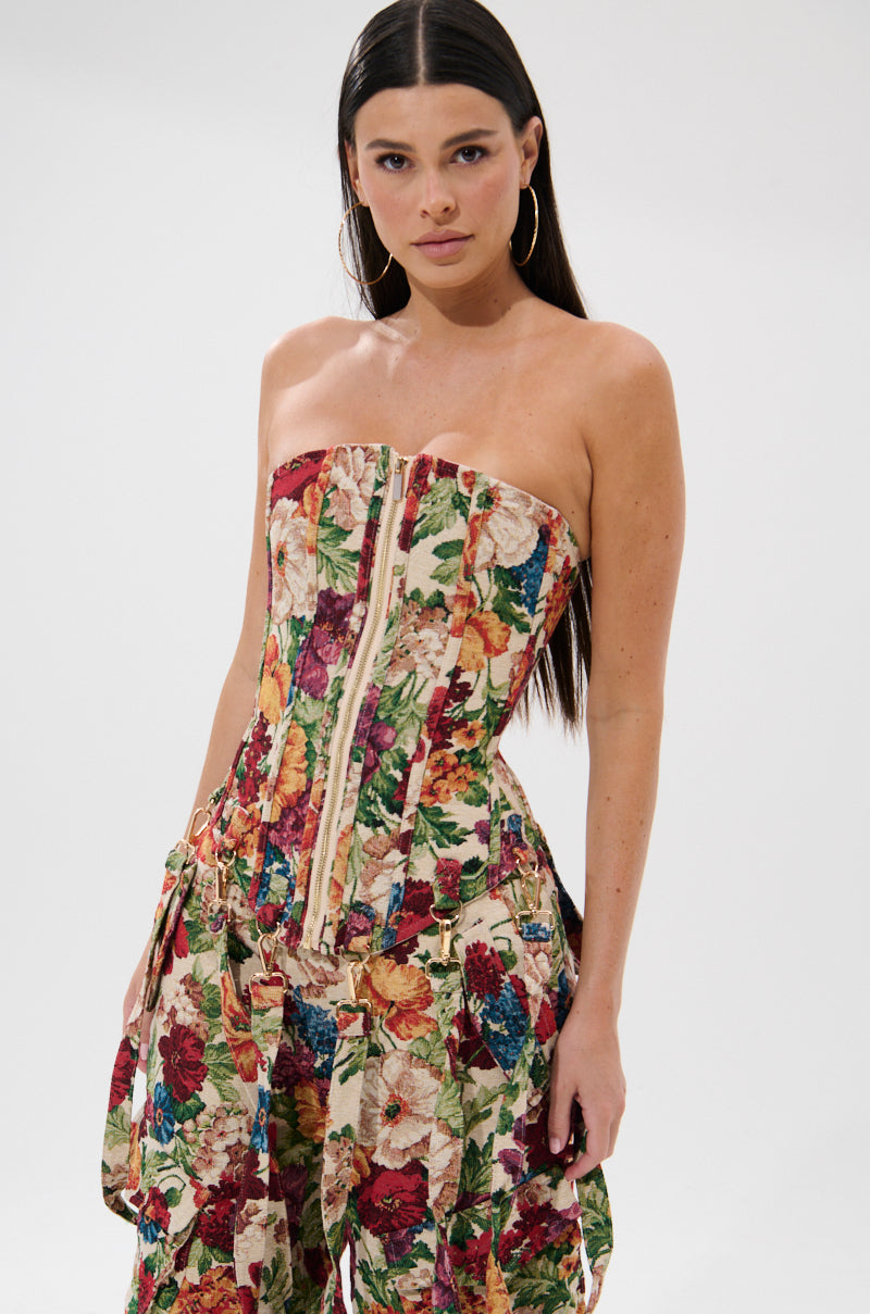 A woman with long dark hair models the FLORAL FOR DAYS BROCADE CORSET and matching pants, featuring a floral design and detachable garter straps. She stands against a white background, looking confidently at the camera.