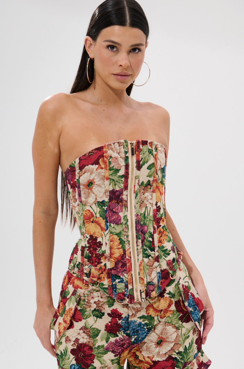 A woman with long dark hair wears the FLORAL FOR DAYS BROCADE CORSET, a strapless, floral brocade top in red, yellow, and green hues with a front zipper and detachable garter straps. She is standing against a white background.