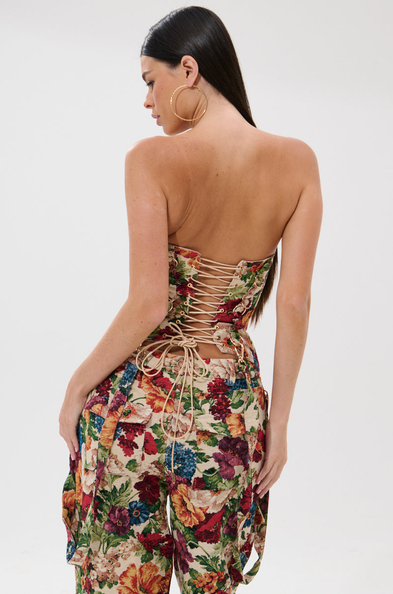 A woman with long dark hair wears large hoop earrings and the FLORAL FOR DAYS BROCADE CORSET, featuring a floral print and lace-up back. She stands against a plain light background, turned slightly to the side and looking down.