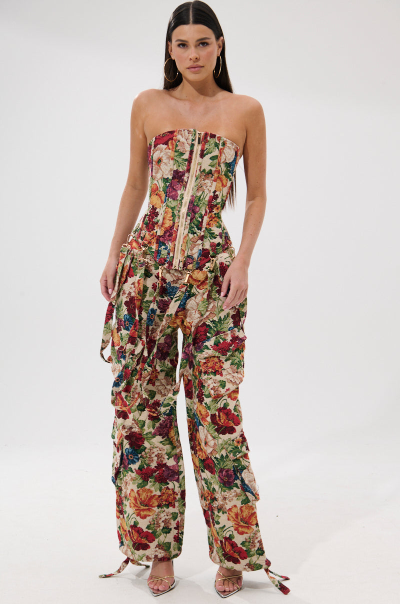 A woman poses against a plain backdrop in the FLORAL FOR DAYS BROCADE CORSET jumpsuit, featuring a strapless, fitted bodice and wide legs with a vibrant floral pattern. She has long, straight dark hair and wears open-toe heels.
