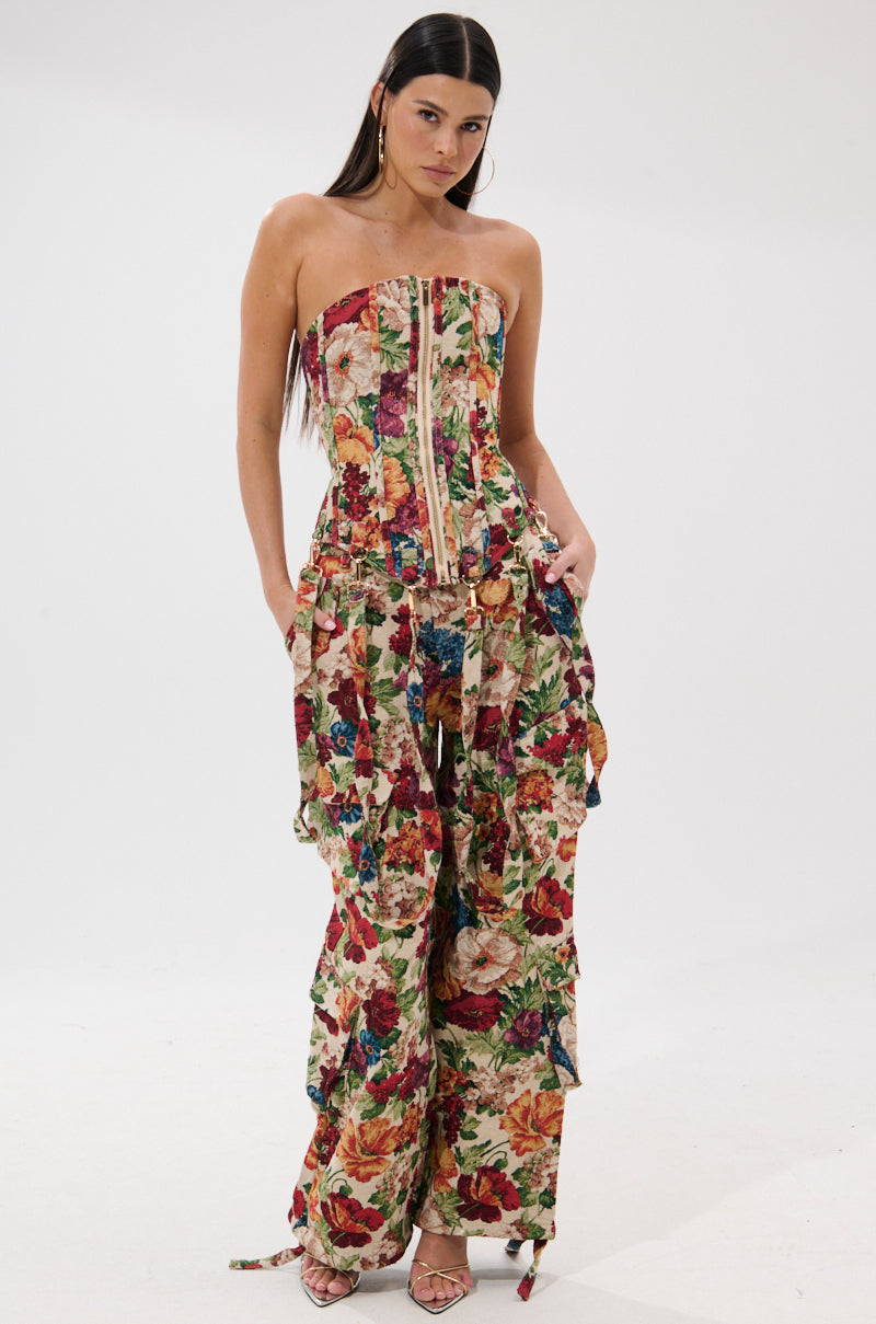 A woman with long, straight dark hair and strappy heels poses against a white background in the FLORAL FOR DAYS BROCADE TROUSER, a high-rise wide-leg pant with a vibrant multicolored floral pattern, ruffled accents, and lace-up front.
