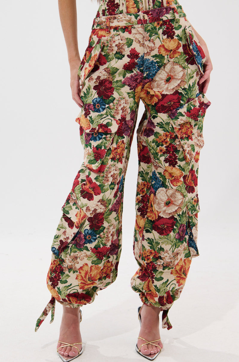 A person wears the FLORAL FOR DAYS BROCADE TROUSER, featuring a high-waisted, wide-leg silhouette with cargo pockets and a vibrant floral print in red, green, yellow, and blue, paired with silver strappy high heels.