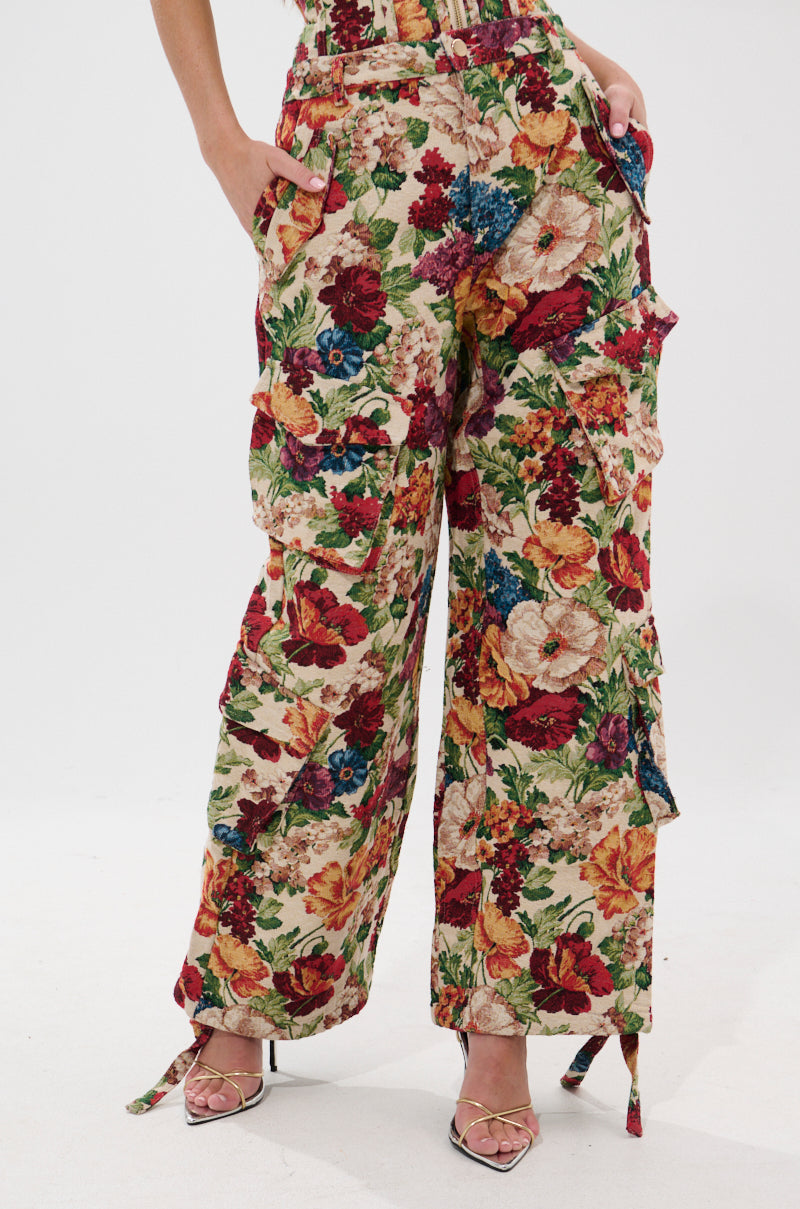 A person models the FLORAL FOR DAYS BROCADE TROUSER, vintage-inspired wide-leg pants featuring a vibrant multicolored floral print, paired with strappy heeled sandals and hands in pockets against a plain white background.