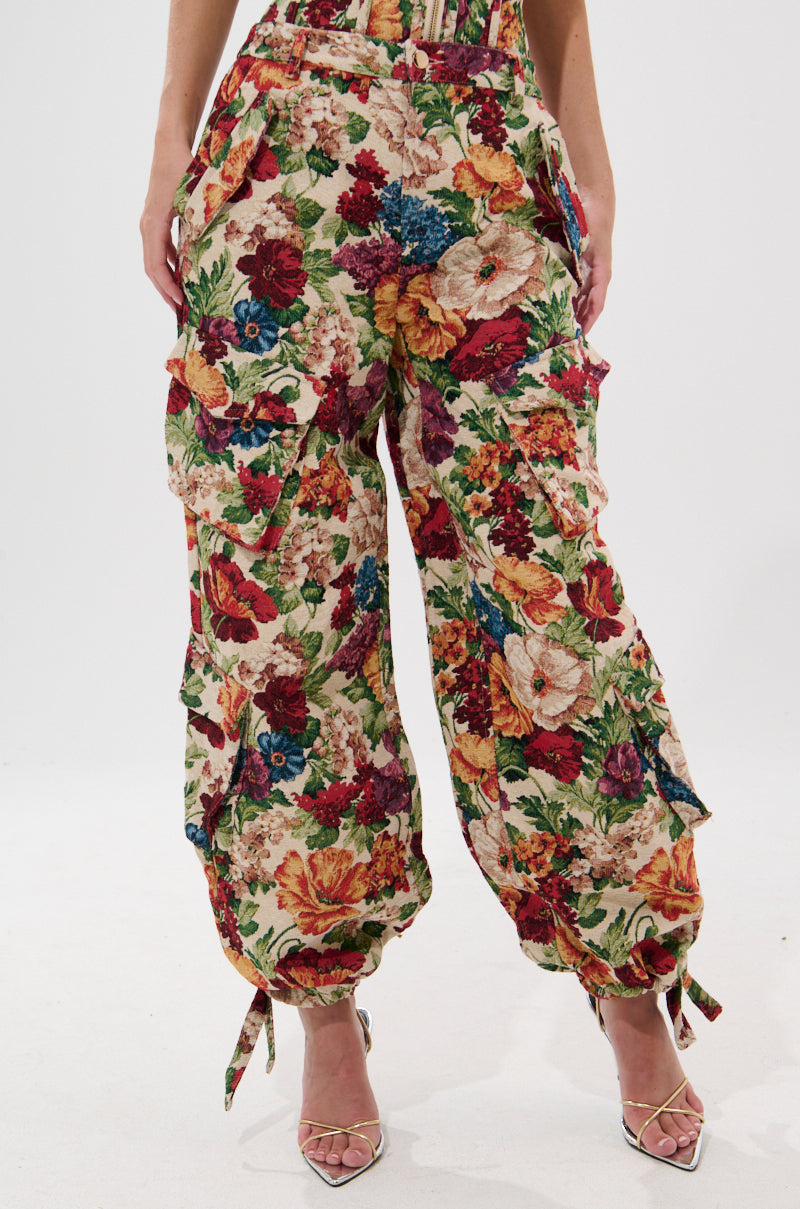 Wearing the FLORAL FOR DAYS BROCADE TROUSER—high-rise, wide-leg pants in a bold floral print with adjustable ankle ties—paired with strappy high-heeled sandals.