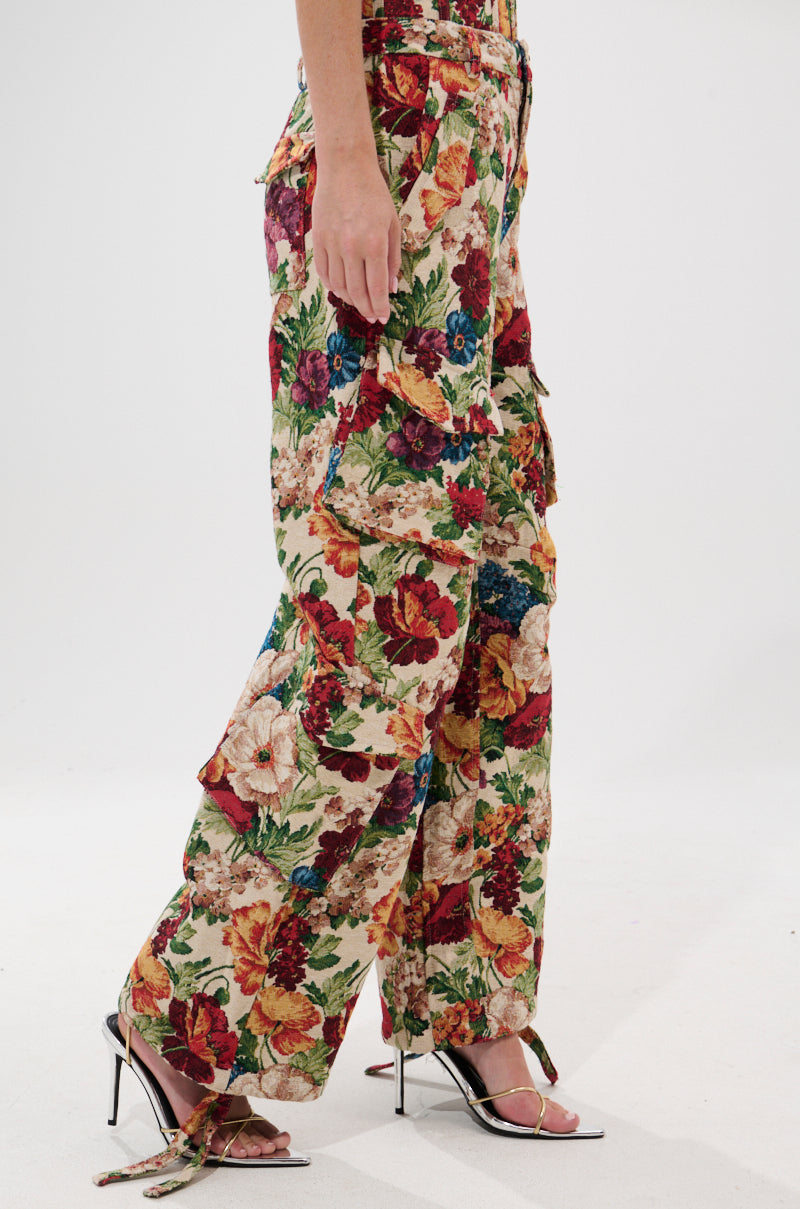 A person stands against a plain white background wearing the FLORAL FOR DAYS BROCADE TROUSER, which features vintage-inspired floral prints and ruffle details, paired with black strappy high-heeled sandals.