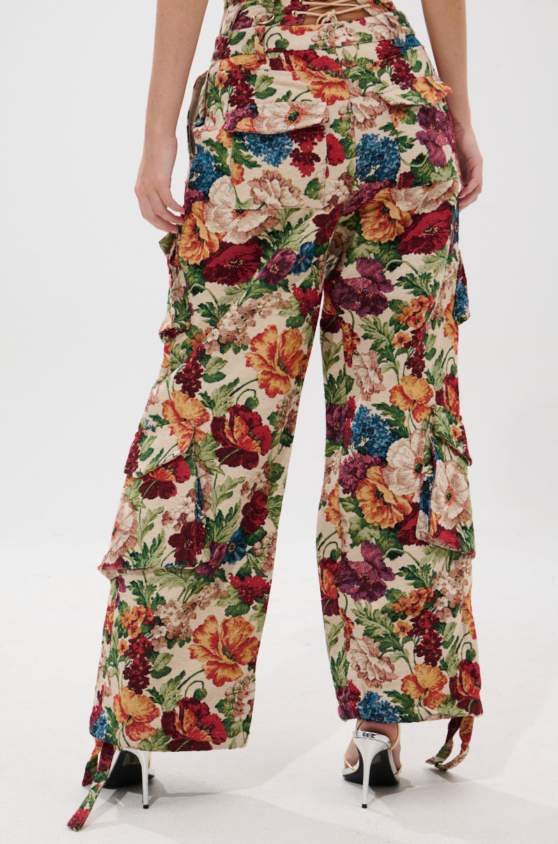 A person faces away wearing the FLORAL FOR DAYS BROCADE TROUSER, featuring a vibrant floral tapestry pattern, wide legs, high-rise fit, and cargo pockets, paired with white heeled sandals. The background is plain white.