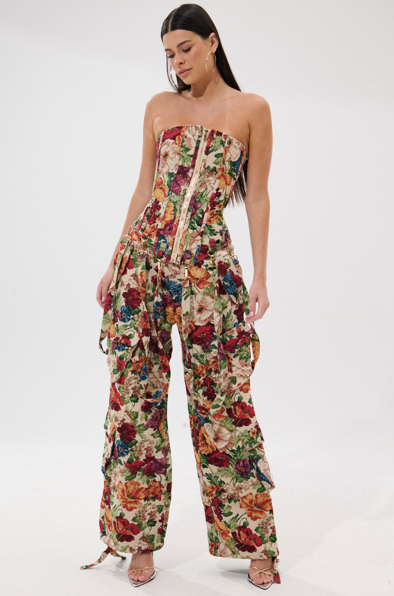 A woman stands against a white background wearing the FLORAL FOR DAYS BROCADE TROUSER—vintage-inspired floral pants with flared legs—paired with a strapless top, strappy heels, and her long dark hair tucked behind her ears.
