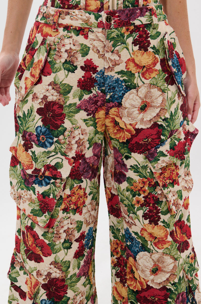 A person wears the FLORAL FOR DAYS BROCADE TROUSER, featuring a vibrant high-rise jacquard design with bold red, yellow, blue, and green floral patterns; their hands rest at their sides.