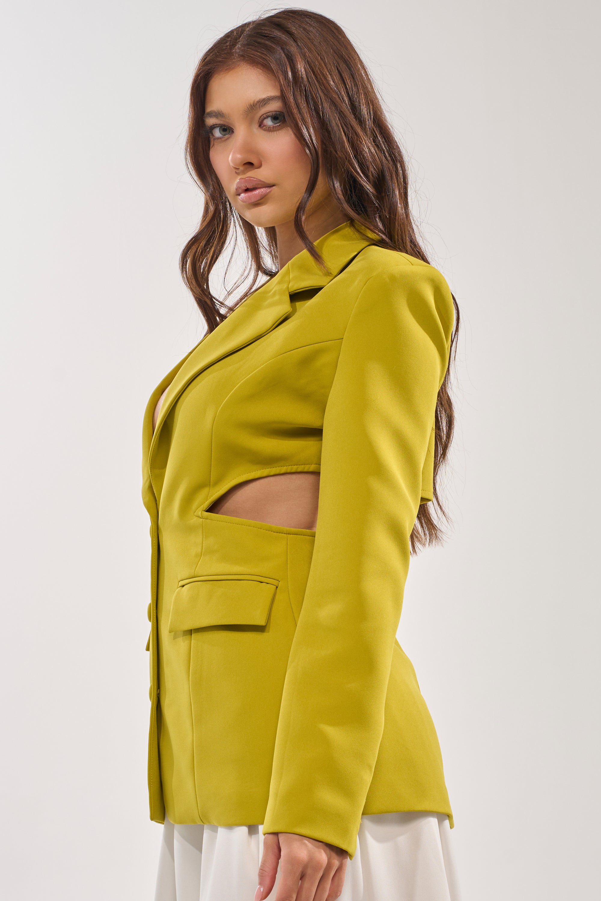 A woman with long brown hair models the PARTY IN THE BACK CUTOUT BLAZER IN CHARTREUSE, styled with a white skirt. She stands sideways against a light background, exuding power dressing flair while looking at the camera. 3