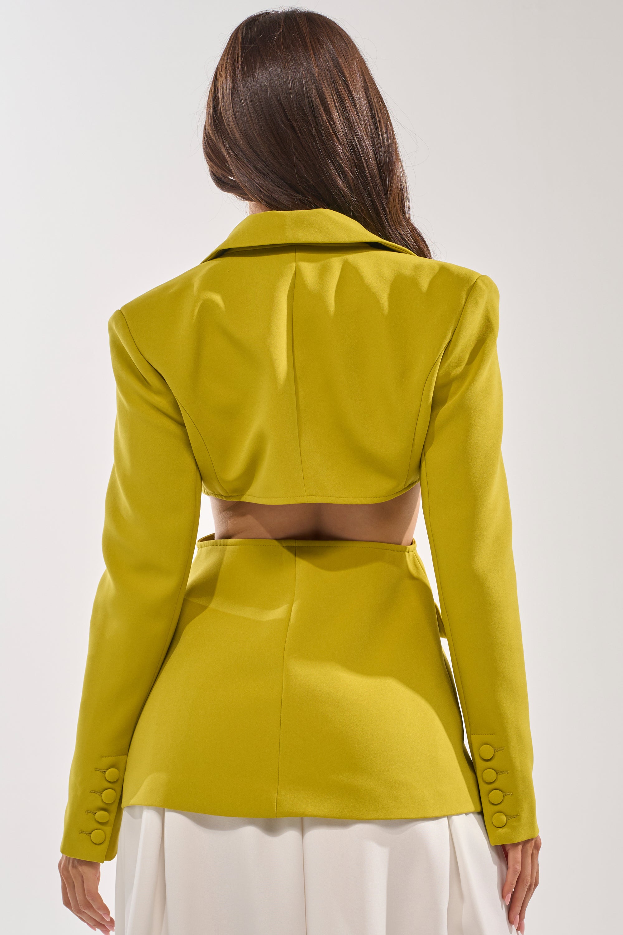 A woman with long brown hair is seen from behind wearing the PARTY IN THE BACK CUTOUT BLAZER IN CHARTREUSE, featuring statement button details and a matching yellow belt over a white pleated skirt. 4