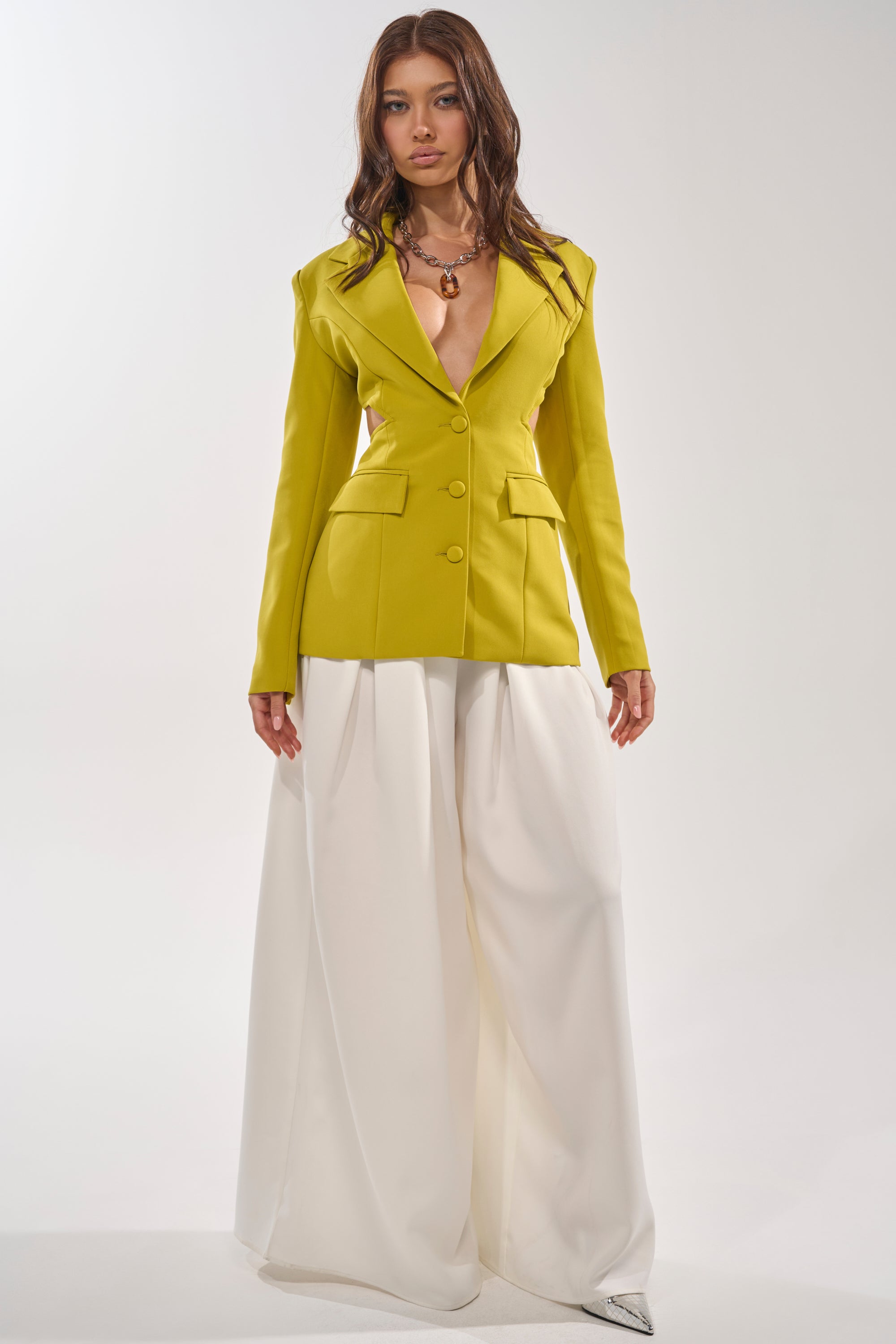 A woman poses against a white background in the PARTY IN THE BACK CUTOUT BLAZER IN CHARTREUSE, styled with wide-legged white trousers, a necklace, pointed shoes, and long wavy brown hair for a modern power look. 5