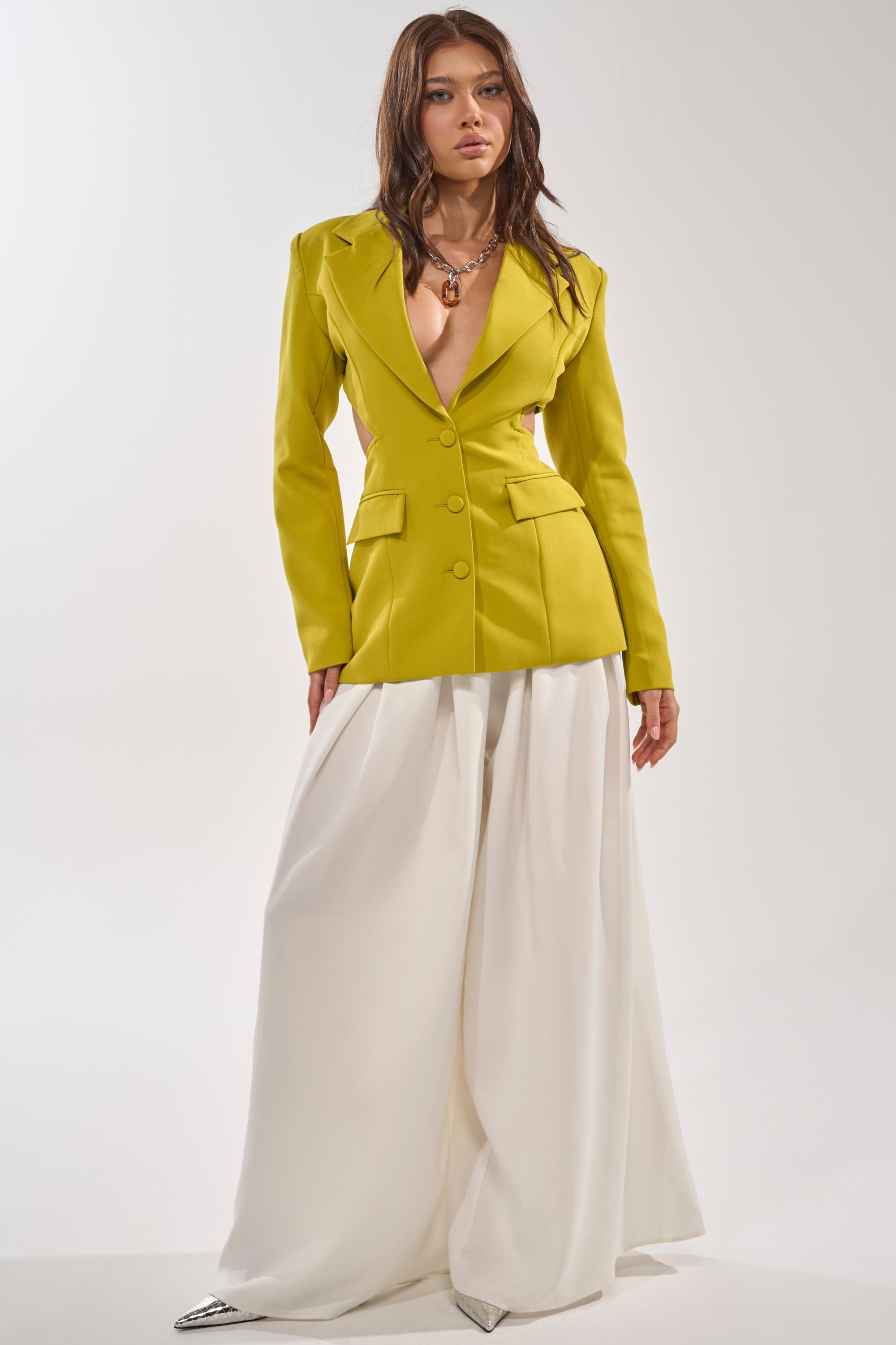 A woman exudes confidence in the PARTY IN THE BACK CUTOUT BLAZER IN CHARTREUSE, styled with wide-legged white pants and layered necklaces—an effortless showcase of modern power dressing against a clean white backdrop. 6