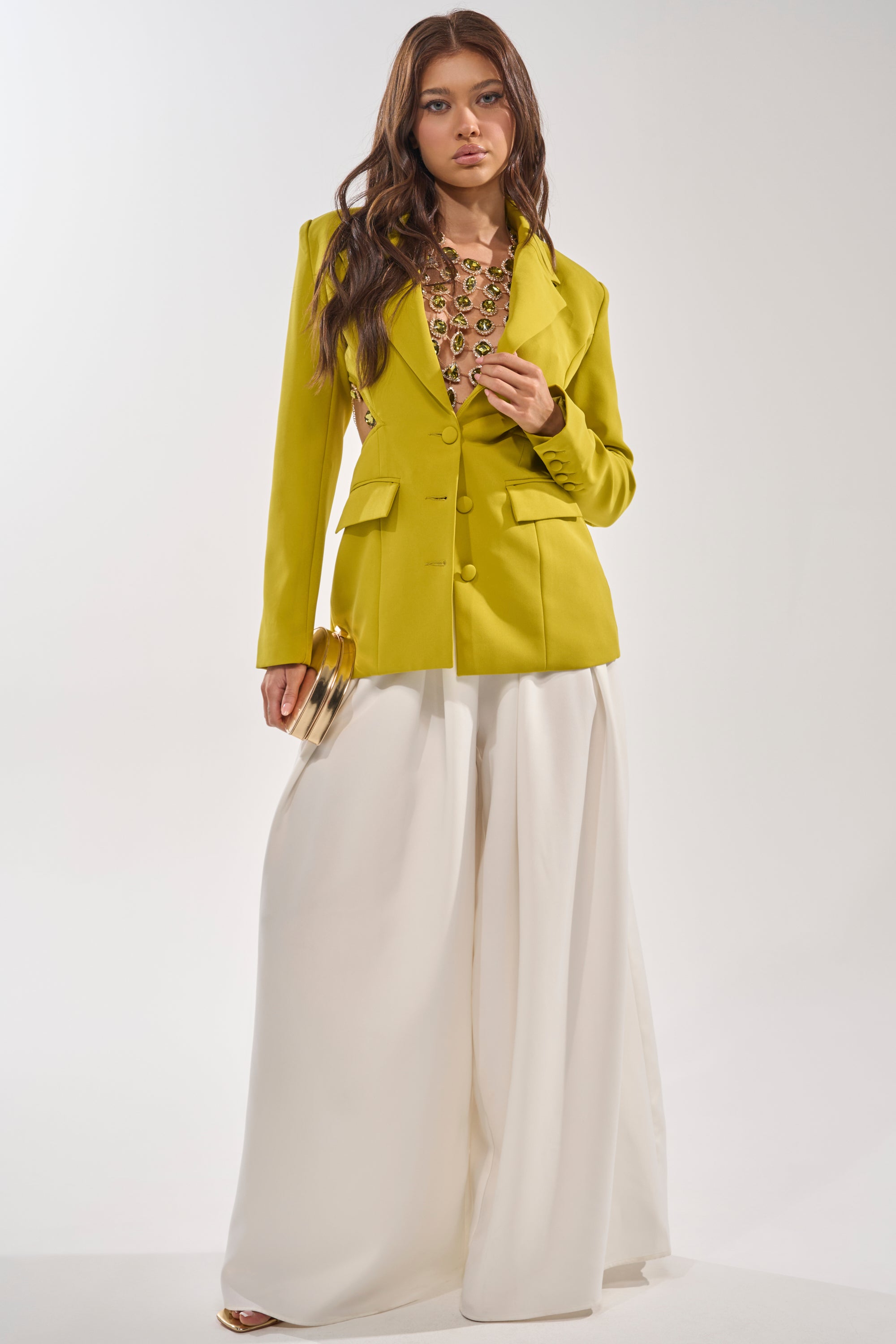 A woman exudes confidence in the PARTY IN THE BACK CUTOUT BLAZER IN CHARTREUSE, styled with a statement necklace, wide-legged white trousers, a gold clutch, and long wavy brown hair against a simple backdrop. 7