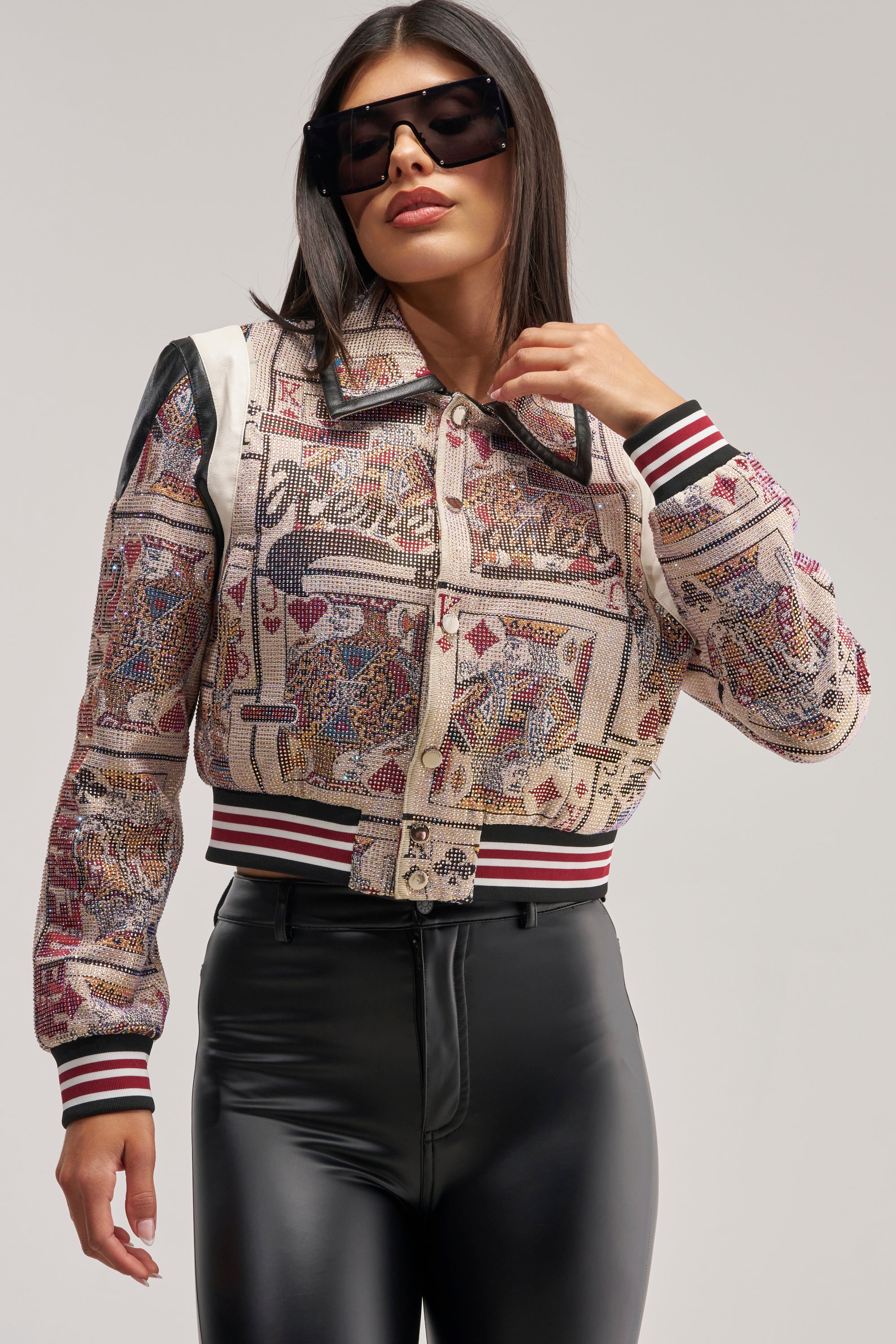 A woman in black sunglasses poses confidently against a plain background, wearing the RAISE YOUR BET RHINESTONE BOMBER—a jacket featuring a playing card graphic and striped cuffs—paired with shiny black high-waisted pants.