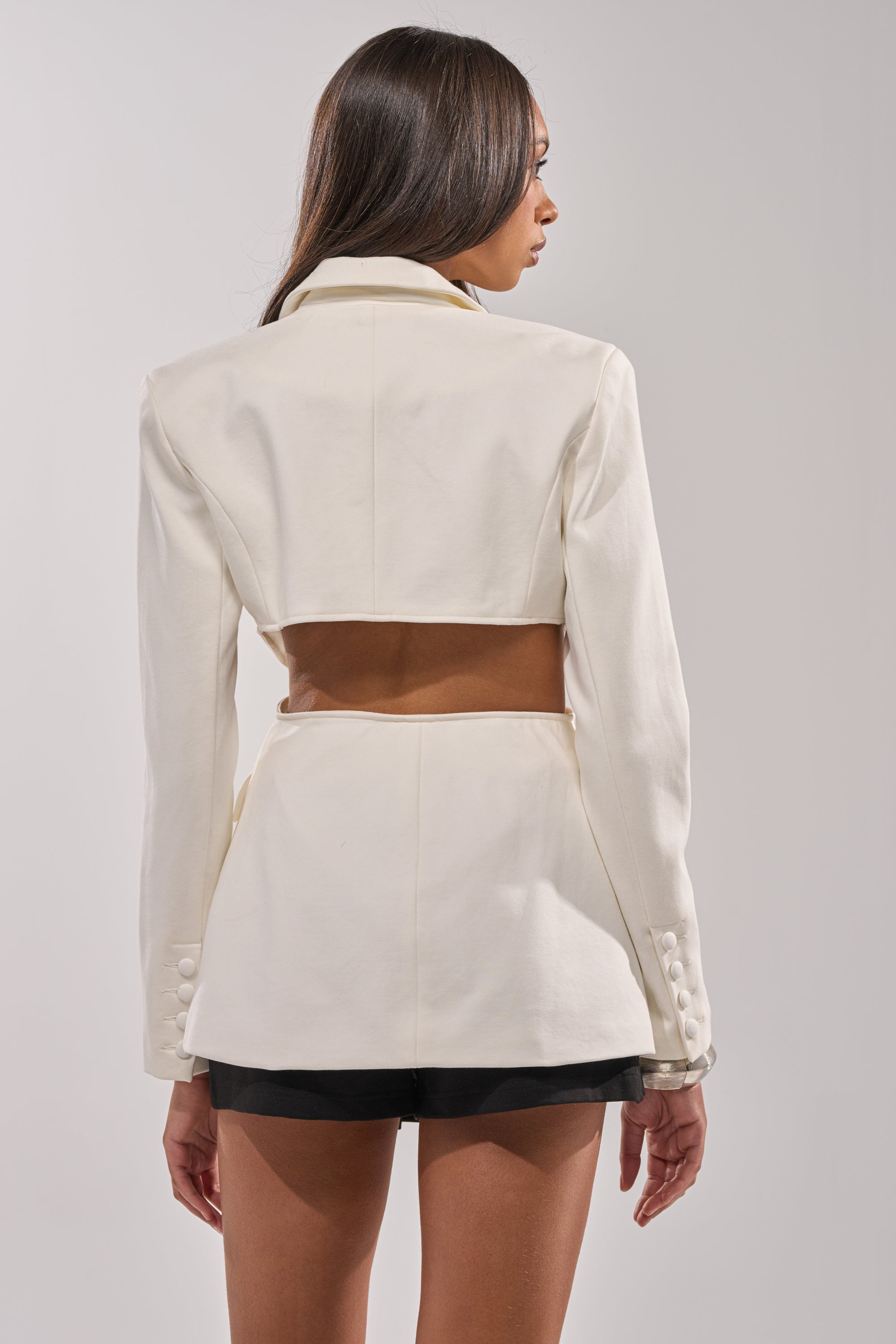 A woman with long brown hair is seen from behind wearing the PARTY IN THE BACK CUTOUT BLAZER IN WHITE, a high-fashion cropped blazer with decorative buttons and long sleeves, styled with black shorts. 4