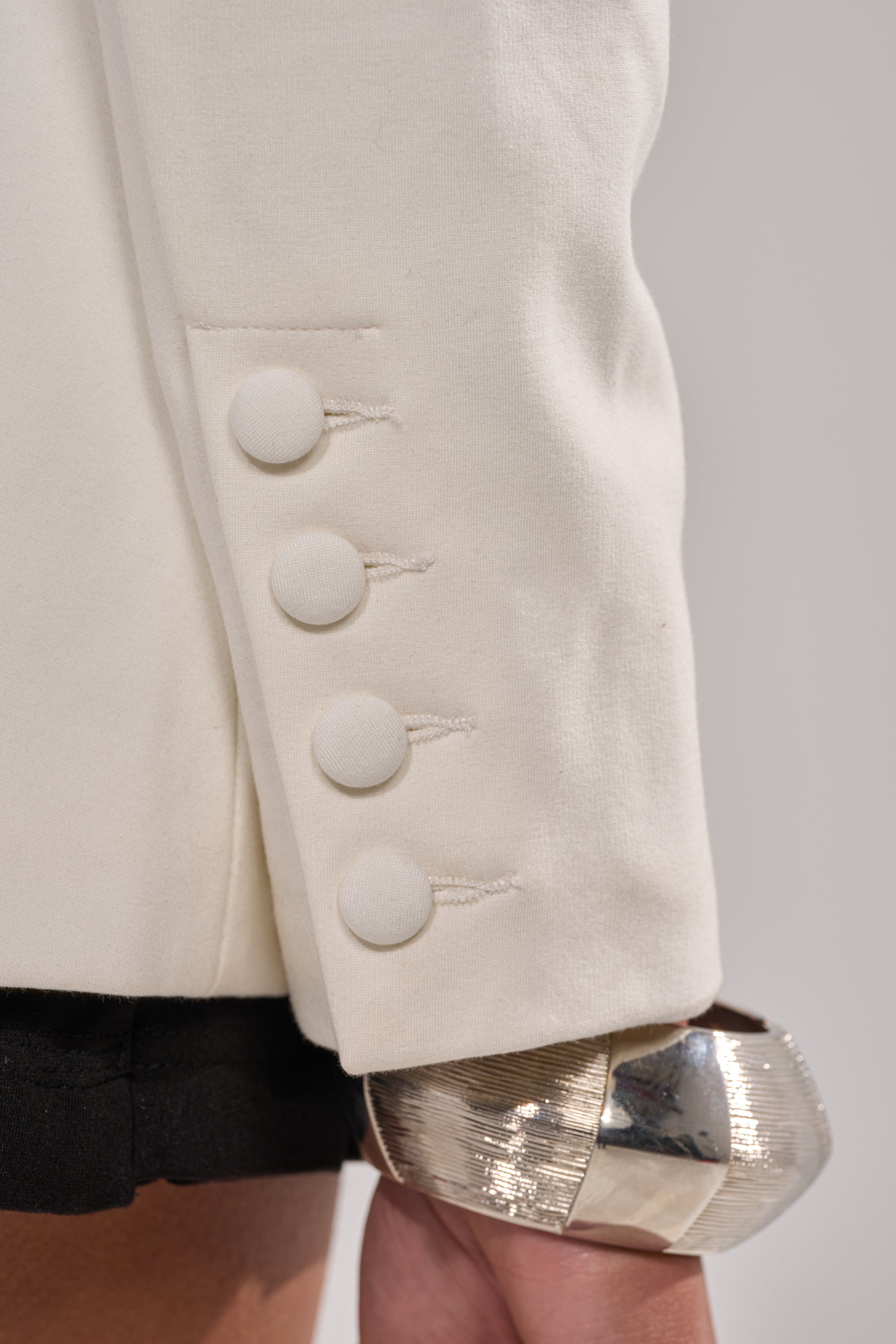 Close-up of the PARTY IN THE BACK CUTOUT BLAZER IN WHITE sleeve, featuring four fabric-covered buttons and visible stitching, worn over a shiny silver cuff bracelet. The background is neutral and blurred. 8
