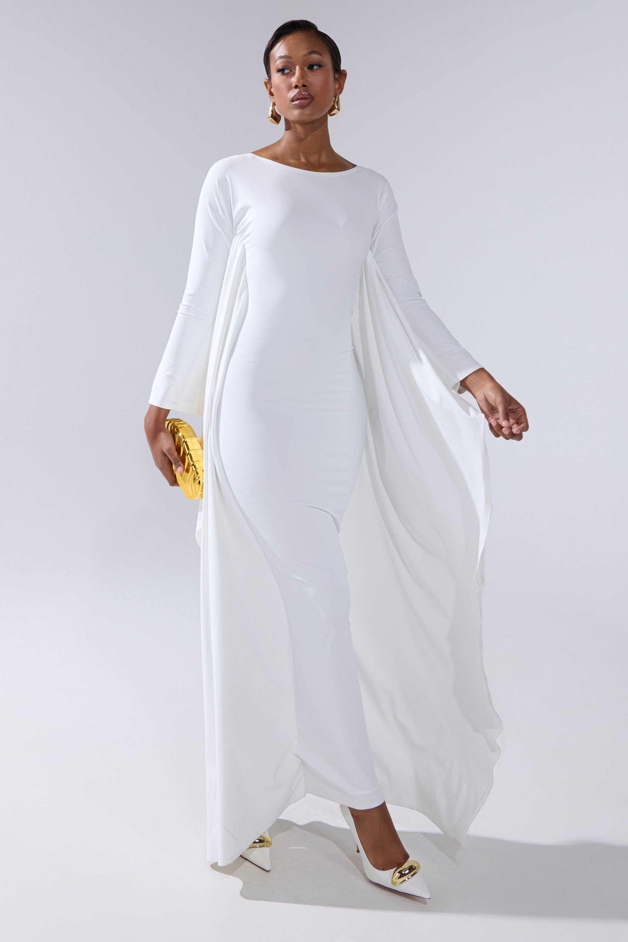 A woman walks confidently in the KYLIE MAXI DRESS WITH RUFFLE SLEEVES IN WHITE, paired with white heels featuring gold accents and a gold clutch, set against a plain light background.