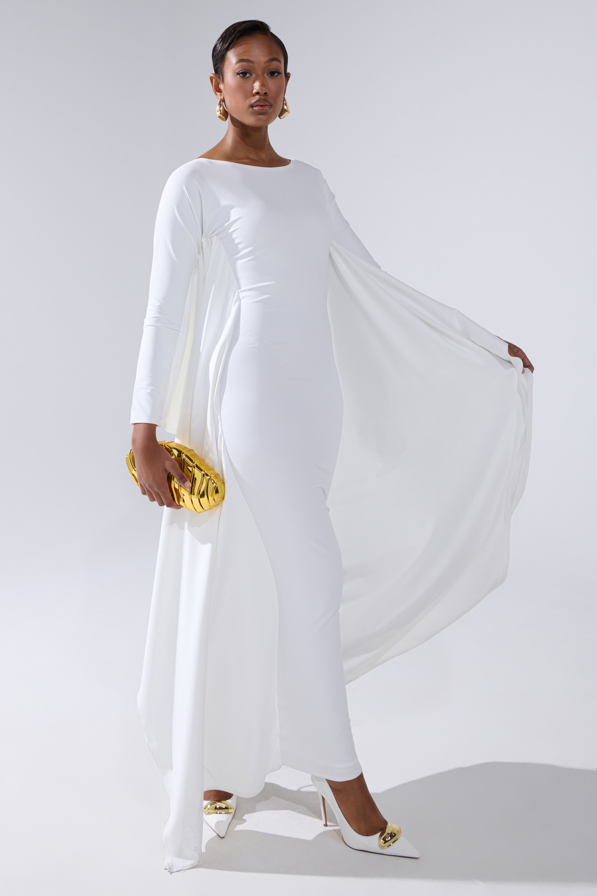 A woman poses confidently in the KYLIE MAXI DRESS WITH RUFFLE SLEEVES IN WHITE, paired with a gold clutch and white heels with gold accents, against a plain light background.