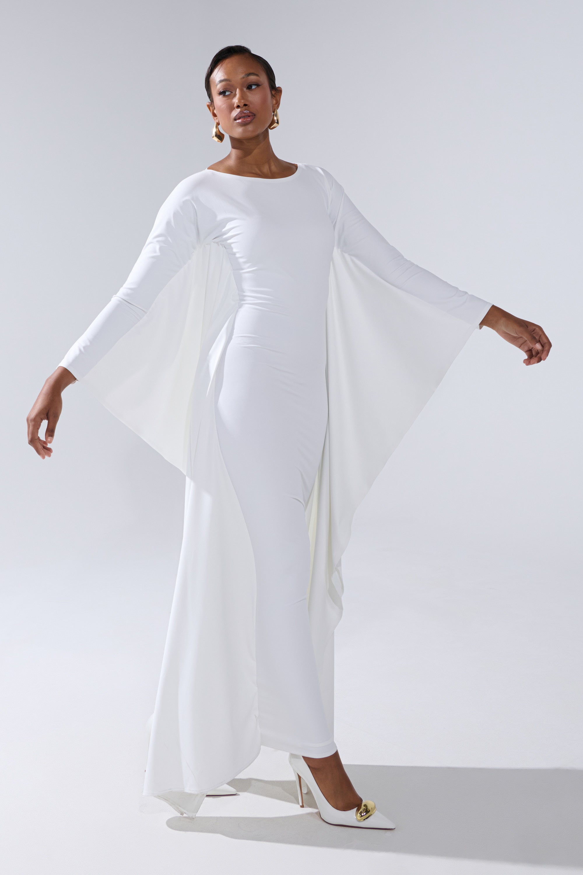 A woman poses gracefully in the KYLIE MAXI DRESS WITH RUFFLE SLEEVES IN WHITE, styled with white heels and gold earrings, her hair neatly pulled back, against a simple, light background.