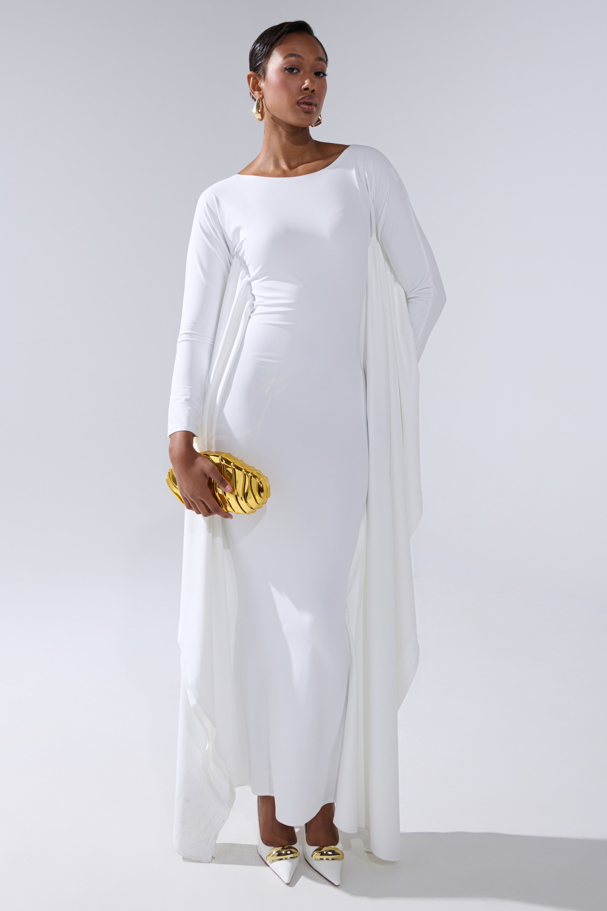 A woman stands confidently in the KYLIE MAXI DRESS WITH RUFFLE SLEEVES IN WHITE, styled with gold earrings, a shiny gold clutch, and pointed white heels with gold tips against a plain light background.