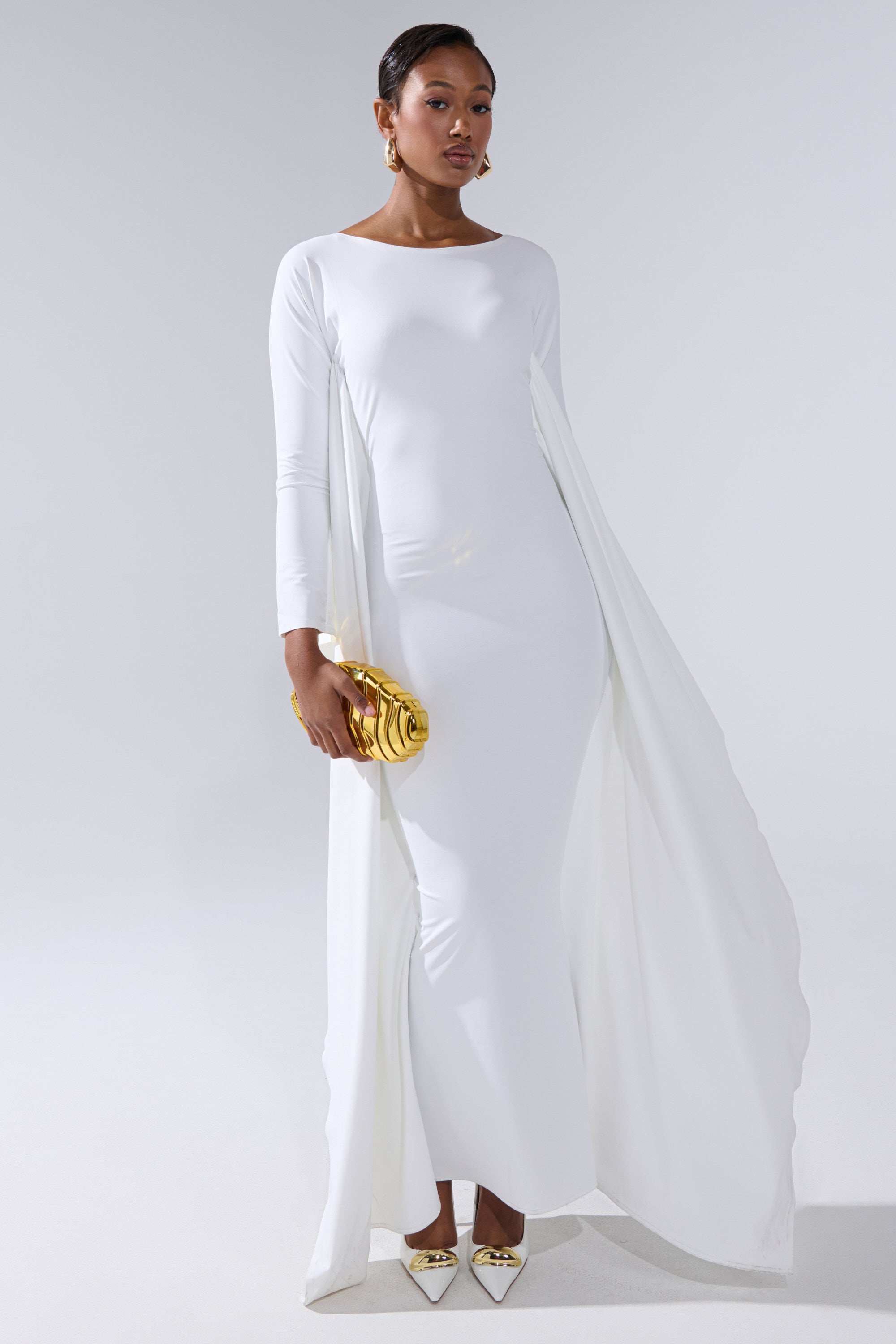 A woman in a white KYLIE MAXI DRESS WITH RUFFLE SLEEVES stands holding a gold clutch. She pairs the look with gold earrings and pointed gold heels, her hair styled back, posing against a plain light background.