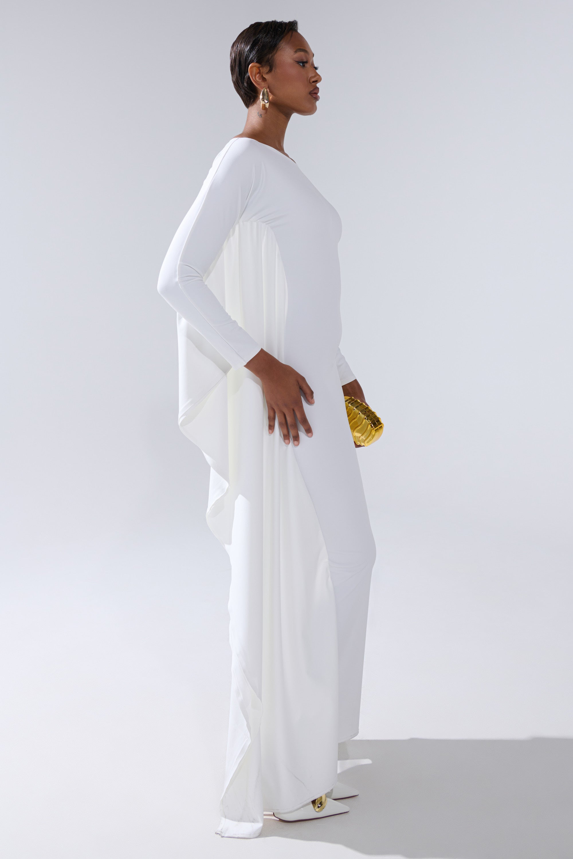 A woman models the KYLIE MAXI DRESS WITH RUFFLE SLEEVES IN WHITE, featuring a cape-like back. She accessorizes with statement earrings and a gold clutch, standing against a simple light background.