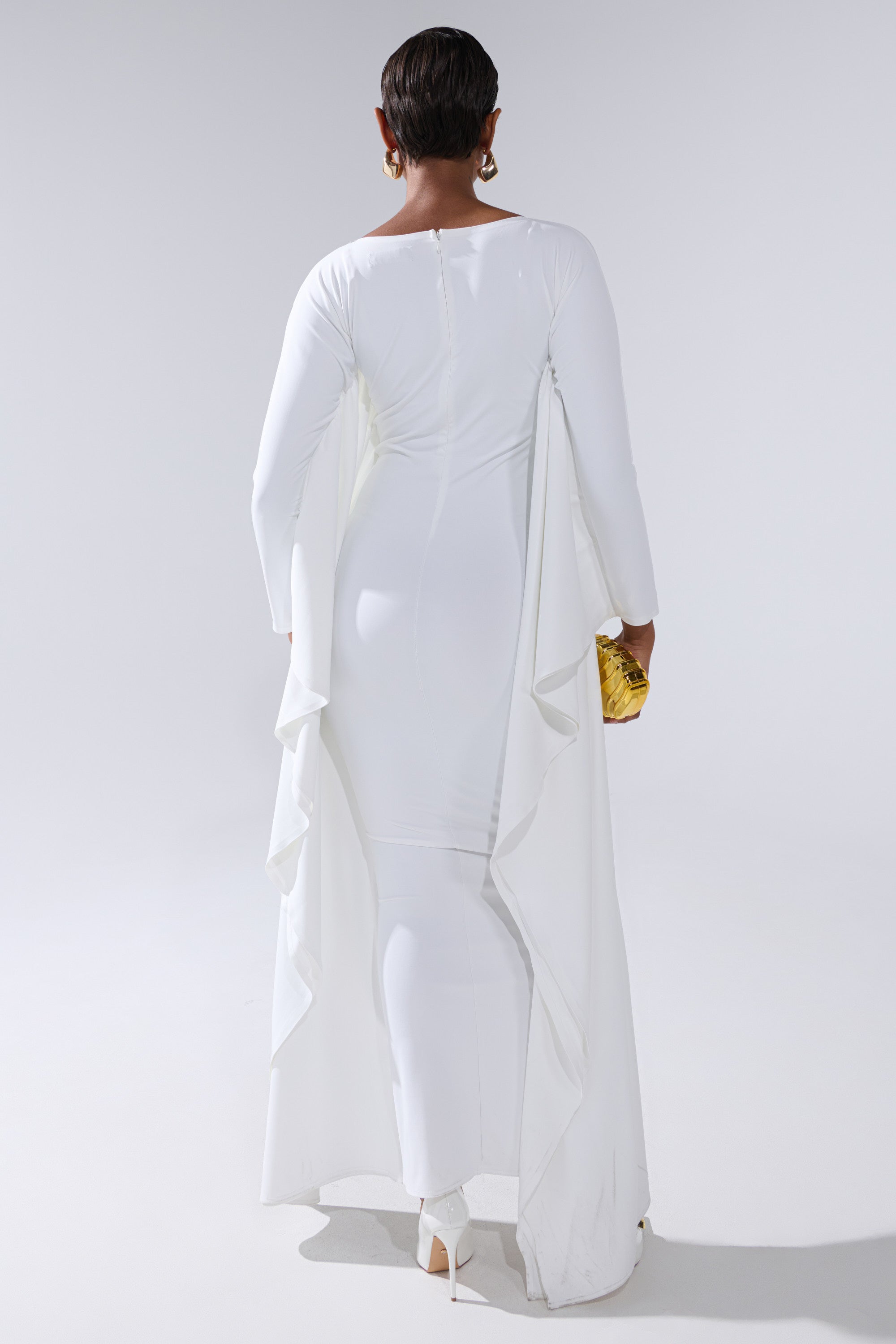 A person with short hair and gold earrings stands facing away, wearing a white KYLIE MAXI DRESS WITH RUFFLE SLEEVES and holding a small yellow clutch against a plain, light-colored background.
