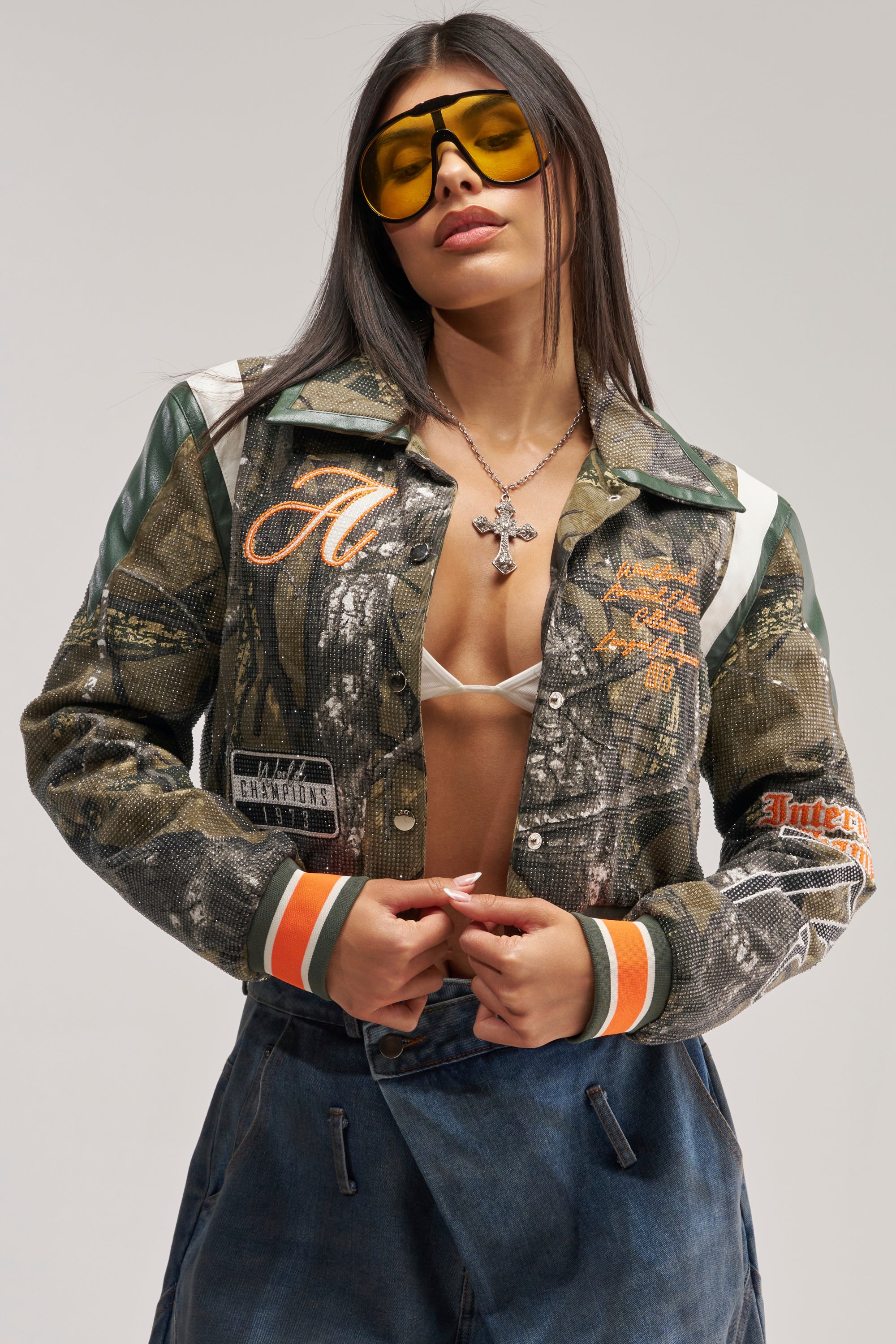 A woman poses confidently with her hands on her TAPPED IN RHINESTONE BOMBER, styled with yellow sunglasses, a white bralette, and blue jeans.