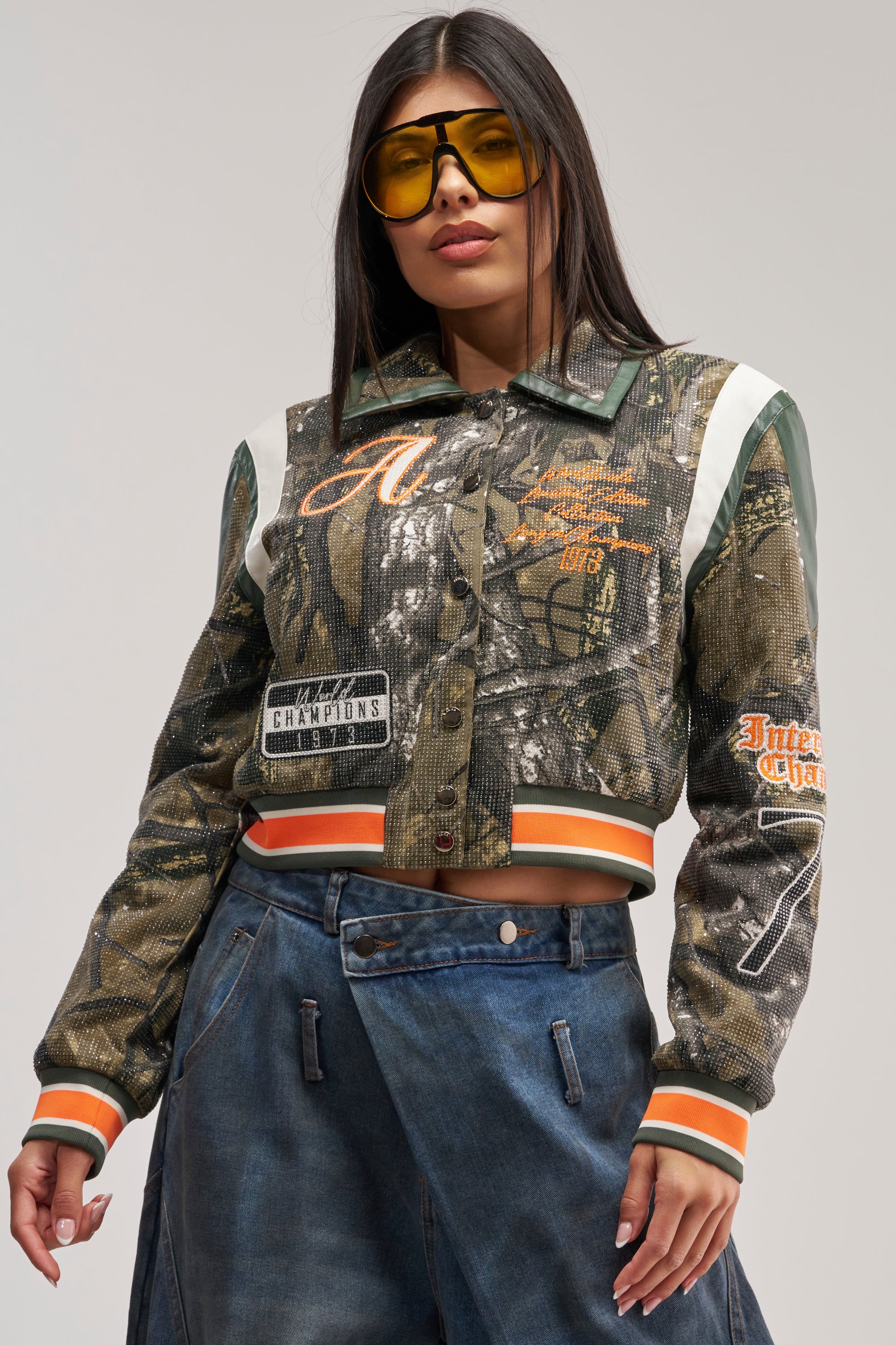 A woman wearing yellow sunglasses and the TAPPED IN RHINESTONE BOMBER—a camo-patterned, cropped streetwear jacket featuring orange and white accents with faux leather trim—paired with high-waisted blue jeans, poses confidently with one hand in her pocket against a plain background.