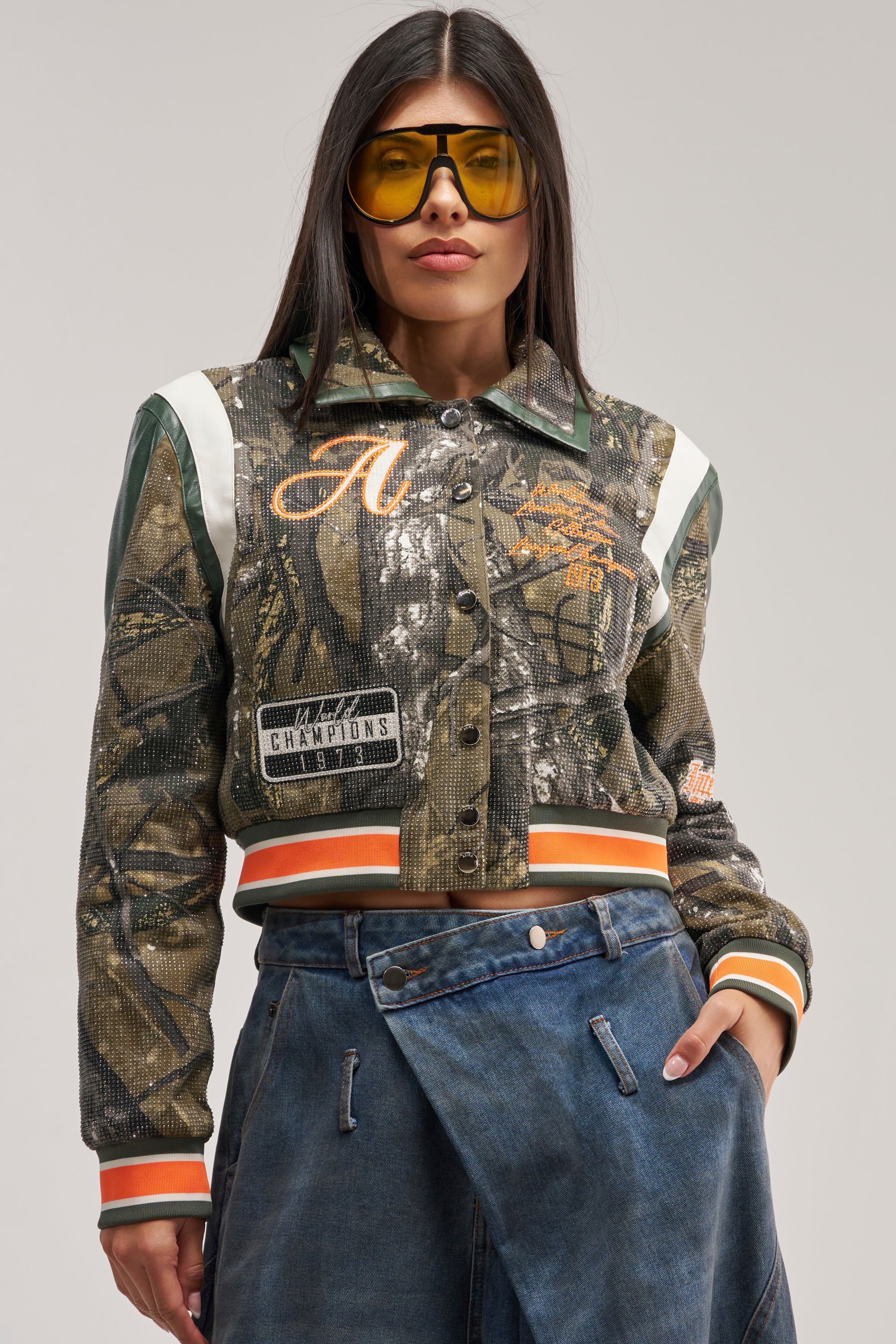 A woman wearing yellow sunglasses and the TAPPED IN RHINESTONE BOMBER—a streetwear jacket with camo print, patches, bold lettering, faux leather trim, and rhinestone details—pairs it with a blue denim skirt as she poses with one hand in her pocket against a neutral background.
