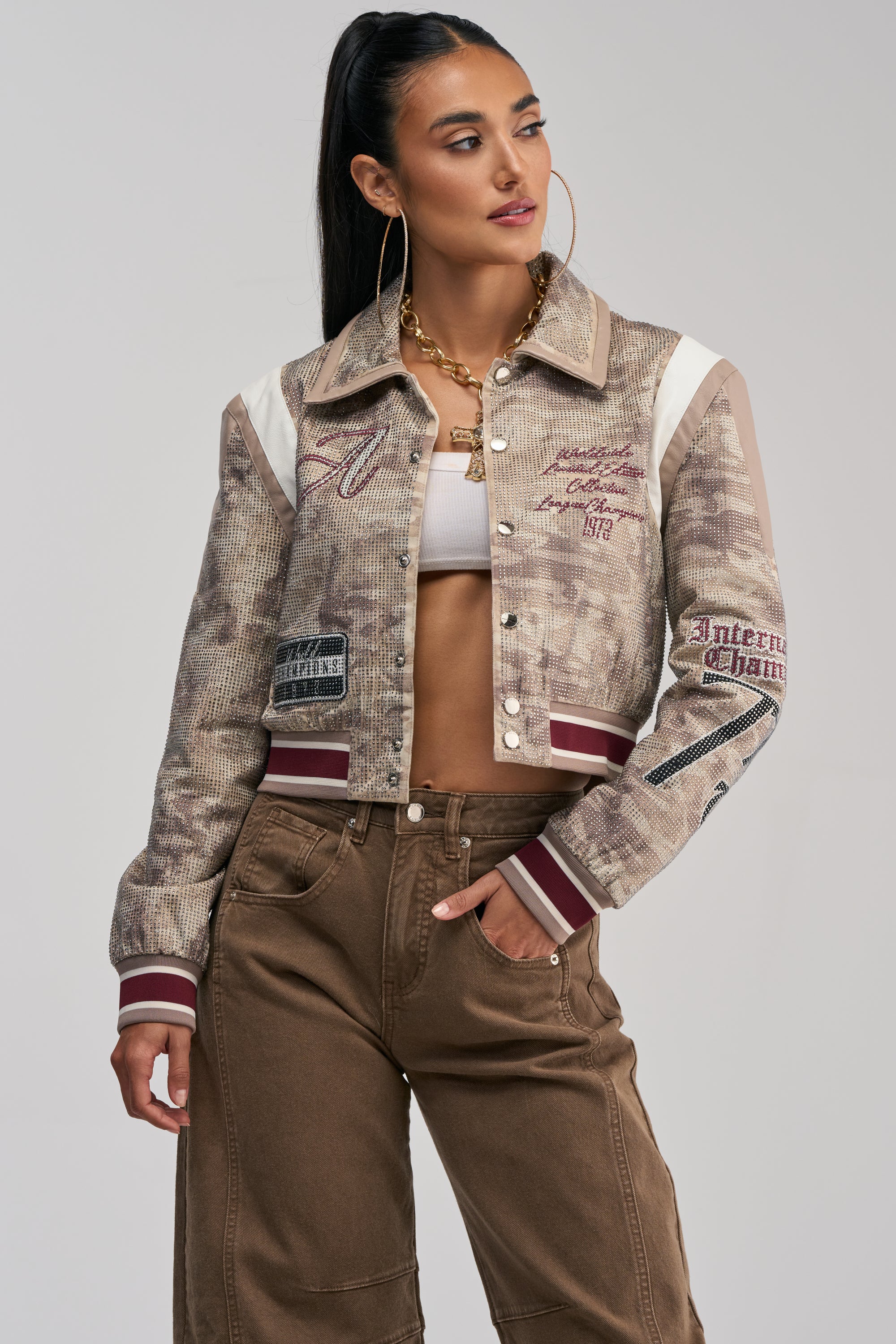 A woman with long dark hair styled in a ponytail wears the BIG EGO RHINESTONE BOMBER IN CAMO, featuring bold embellishments and patches, paired with a white crop top, brown high-waisted pants, and chunky gold jewelry—effortlessly nailing statement street style against a neutral background.